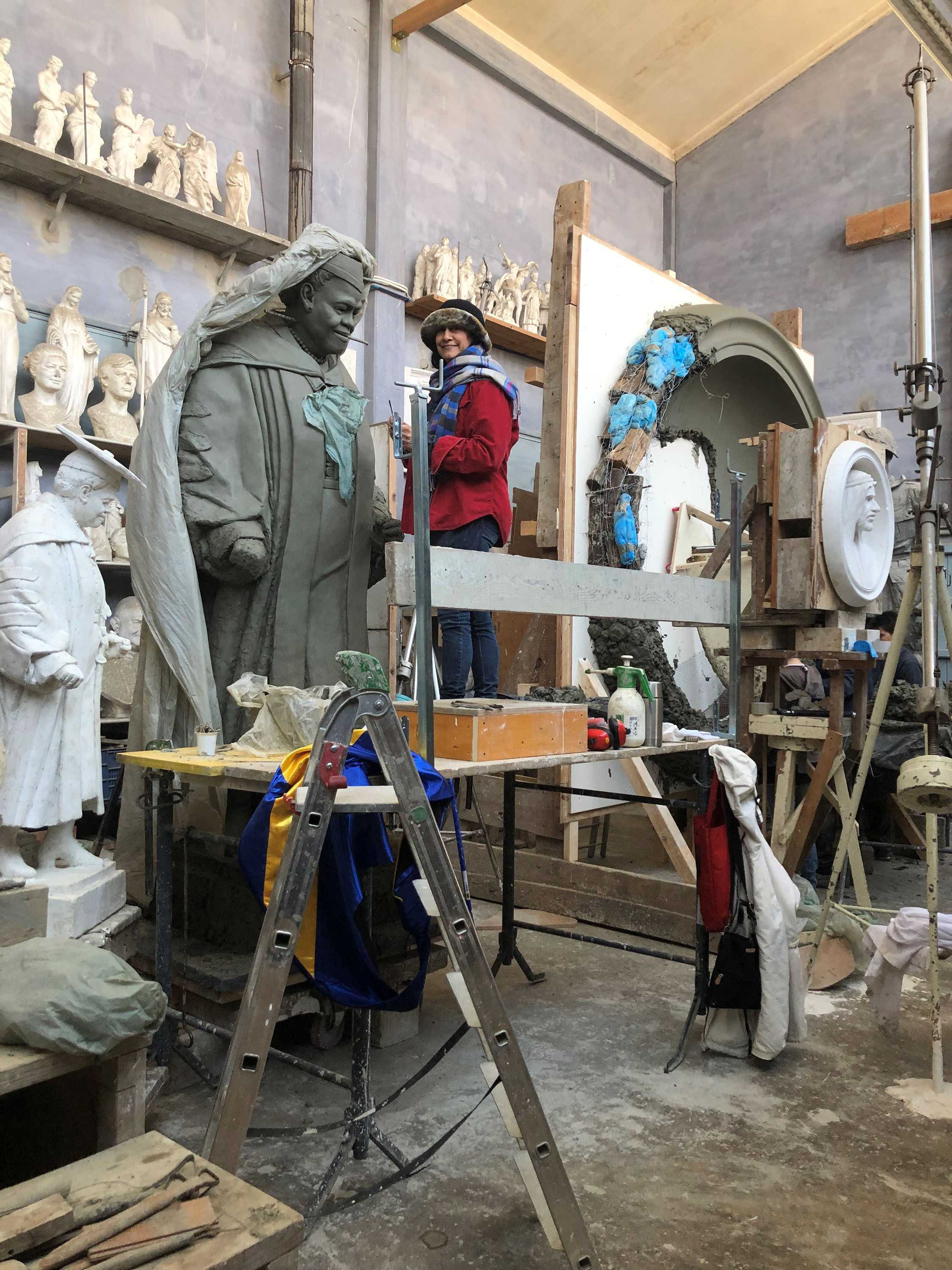 Color photograph showing sculptor's studio with artist working on large clay model.