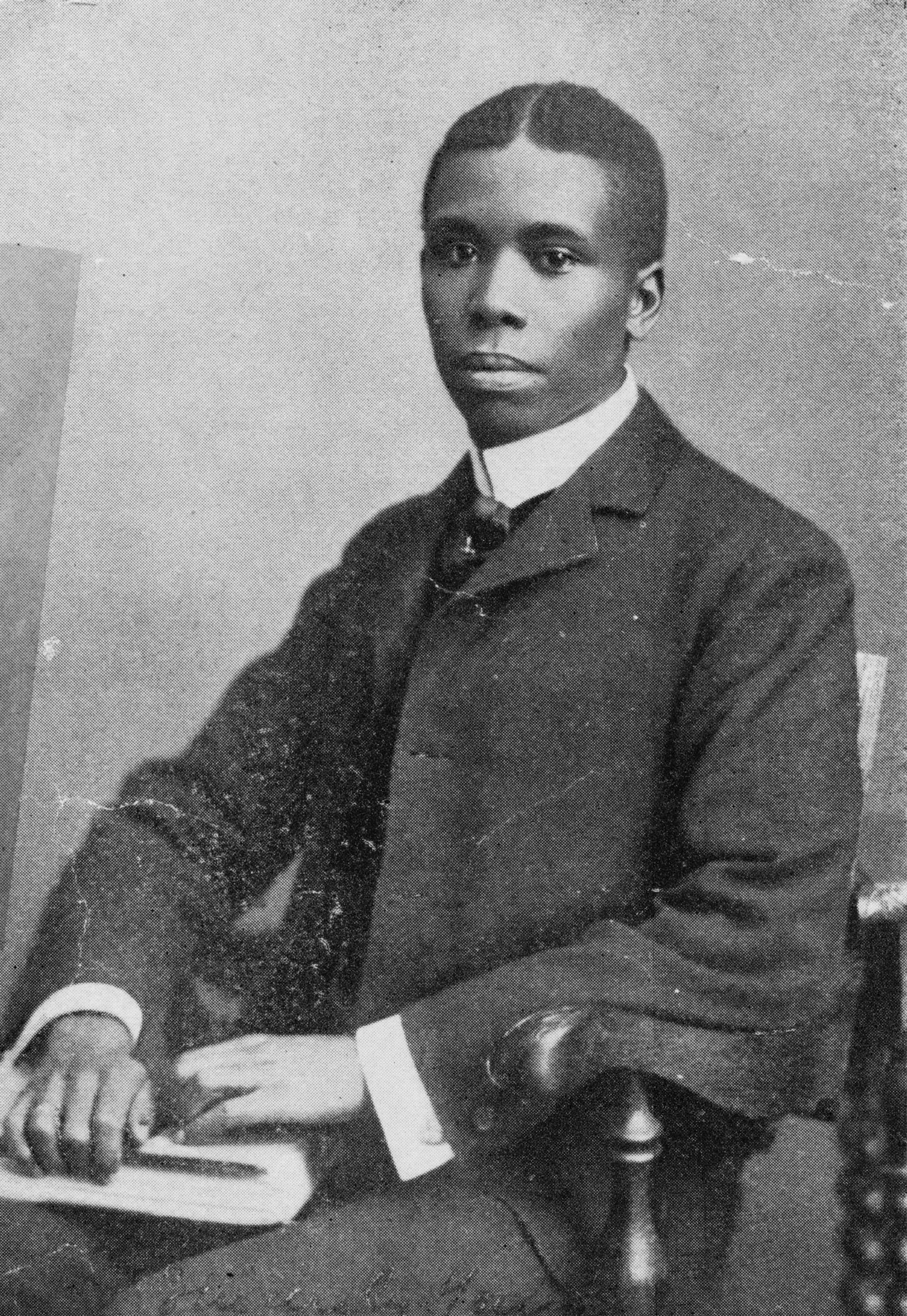 Black and white photograph of young Black man seated in a wooden chair.  He is wearing a suit with a white shirt while holding a book in his lap.