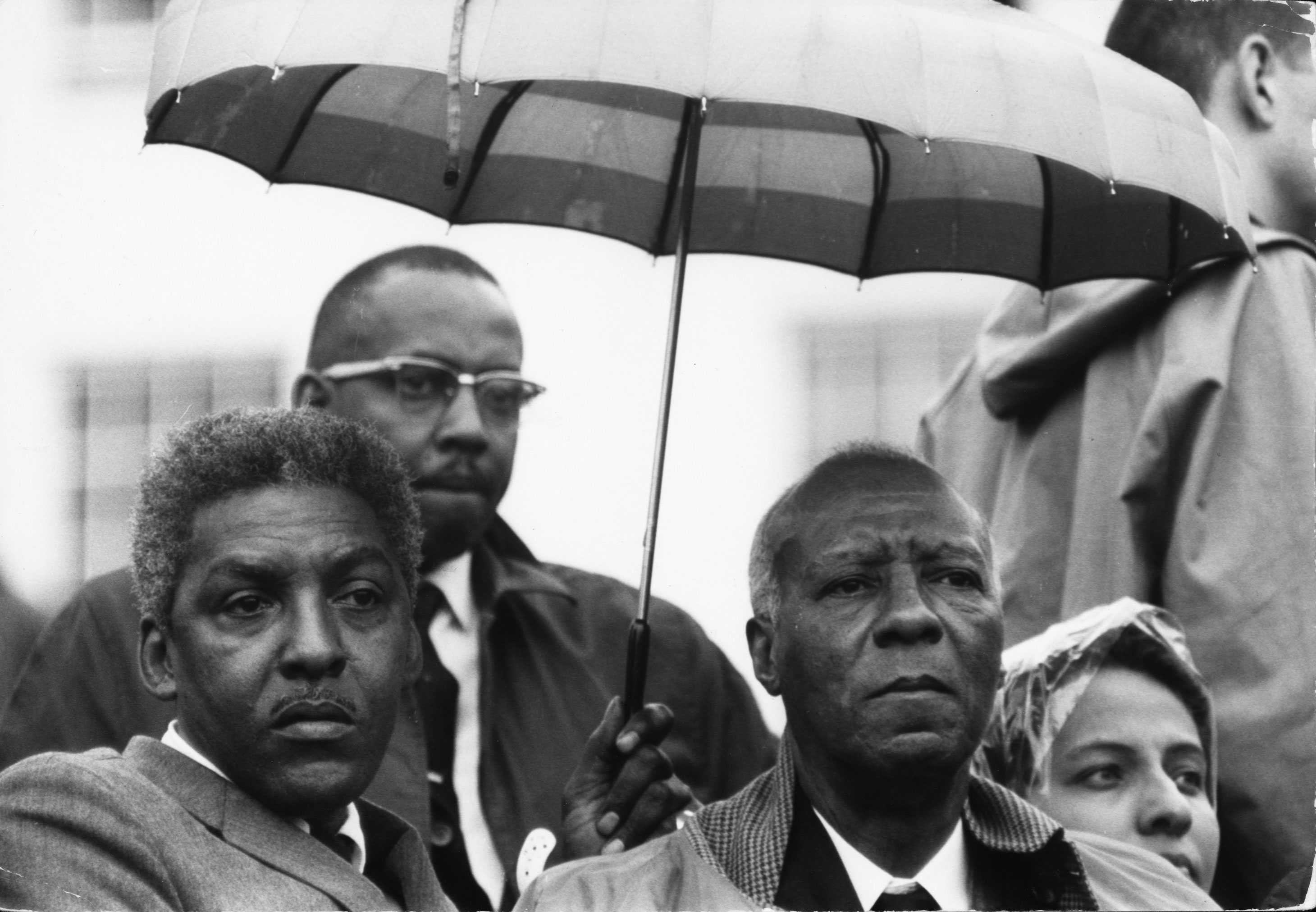 A black-and-white photograph of Bayard Rustin and A. Philip Randolph standing in a crowd with an umbrella over their heads.