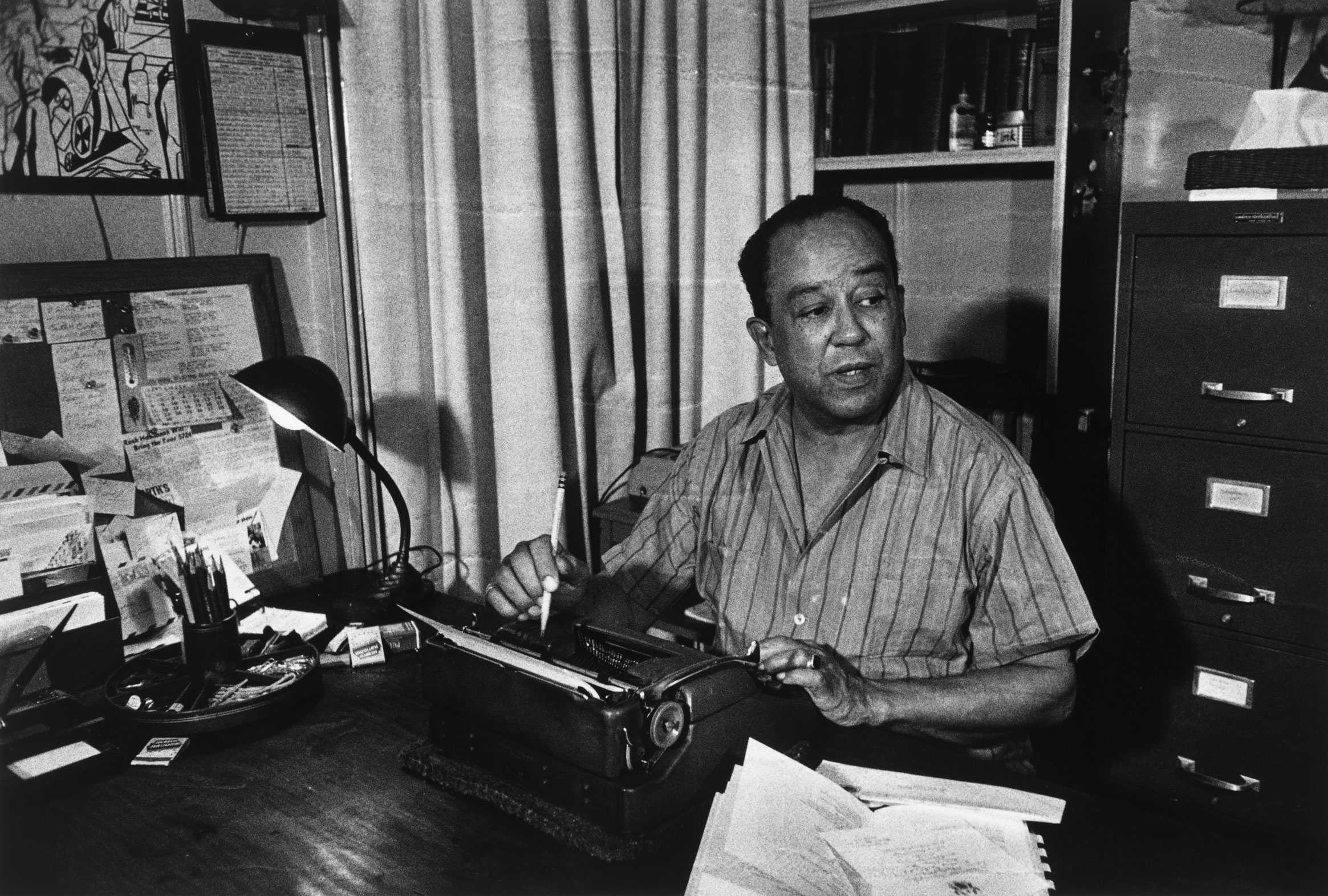 A gelatin silver print of Langston Hughes sitting at a desk with a typewriter, holding a pencil.