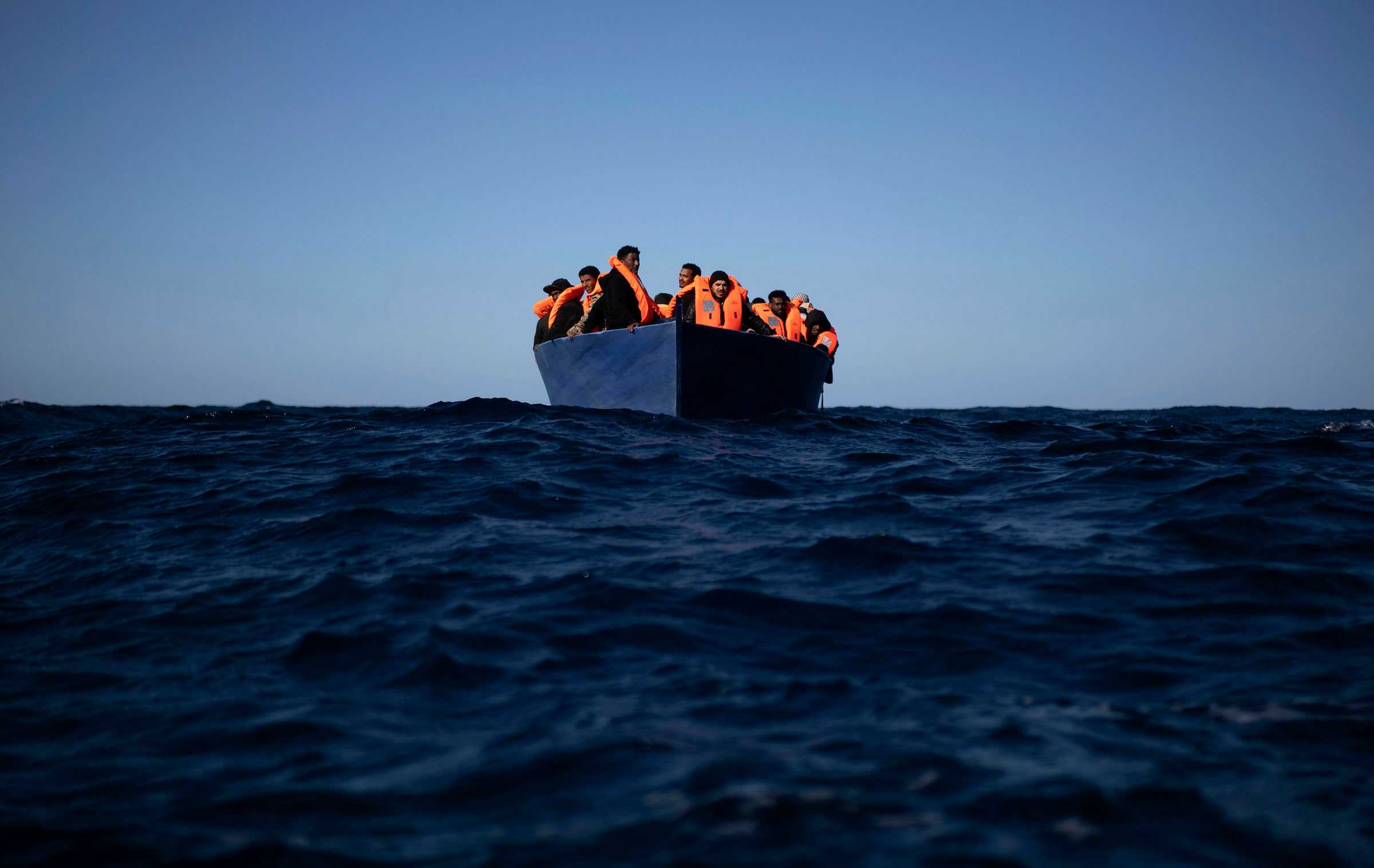 Migrants on a small boat in open water. The migrants are wearing orange life vest.