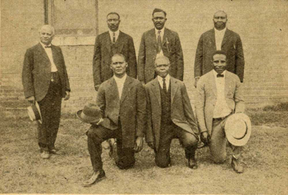 Photograph of 7 men dressed in suits posed in front of a brick building.  Four men stand to the back and three men are kneeling on one knee in the front.