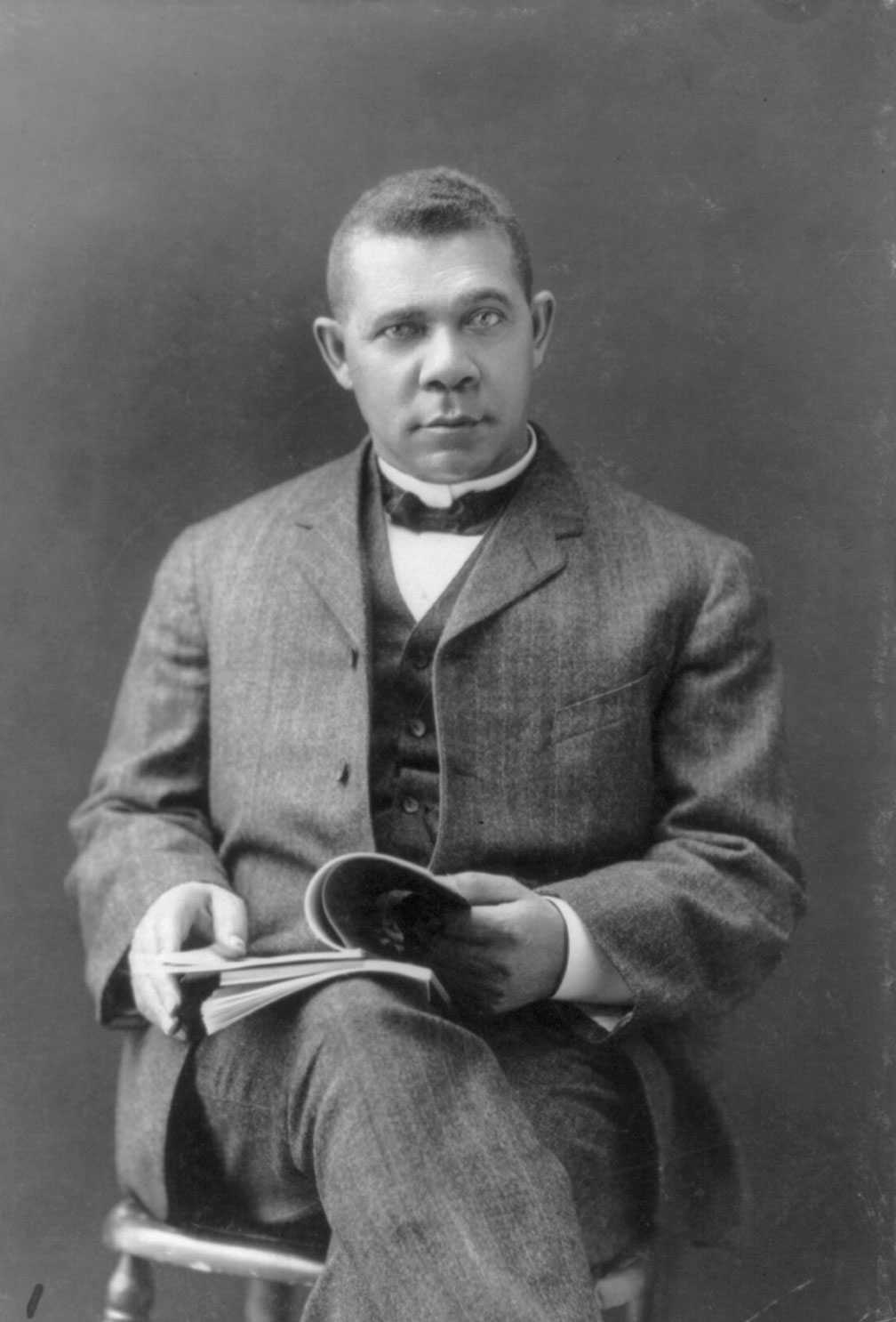 Black and white photograph of African American male seated on a chair, dressed in three-piece suit with a bow tie and holds an open book