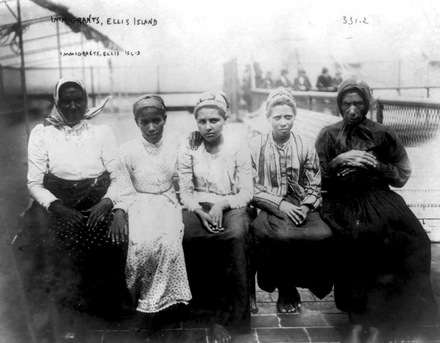 Black and white photograph of 5 females seated on a bench.  They all are wearing headscarves and appear to have bare feet.  There is writing on the image "Immigrants, Ellis Island."