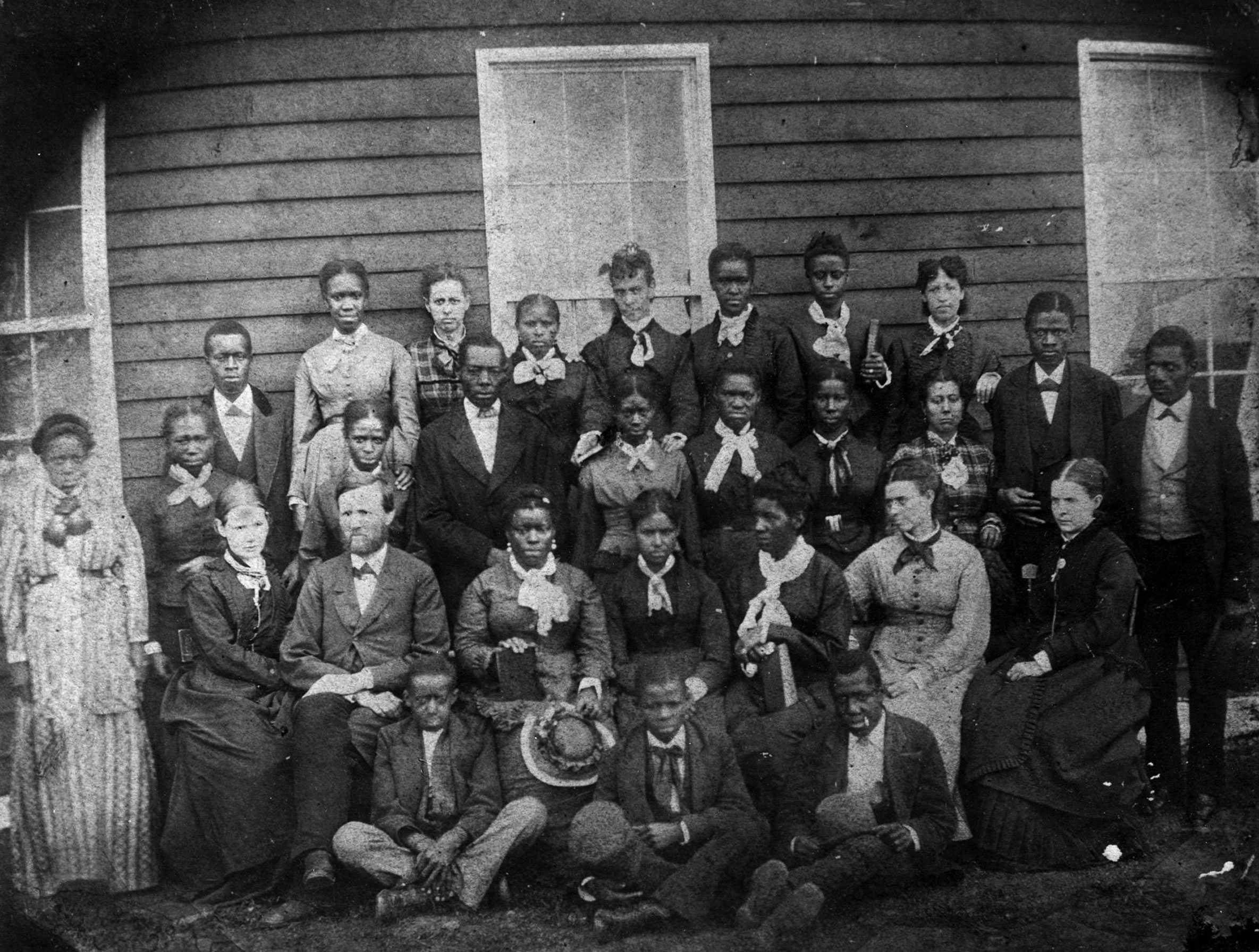 A black and white photo of students and teachers posed outside of their schoolhouse.