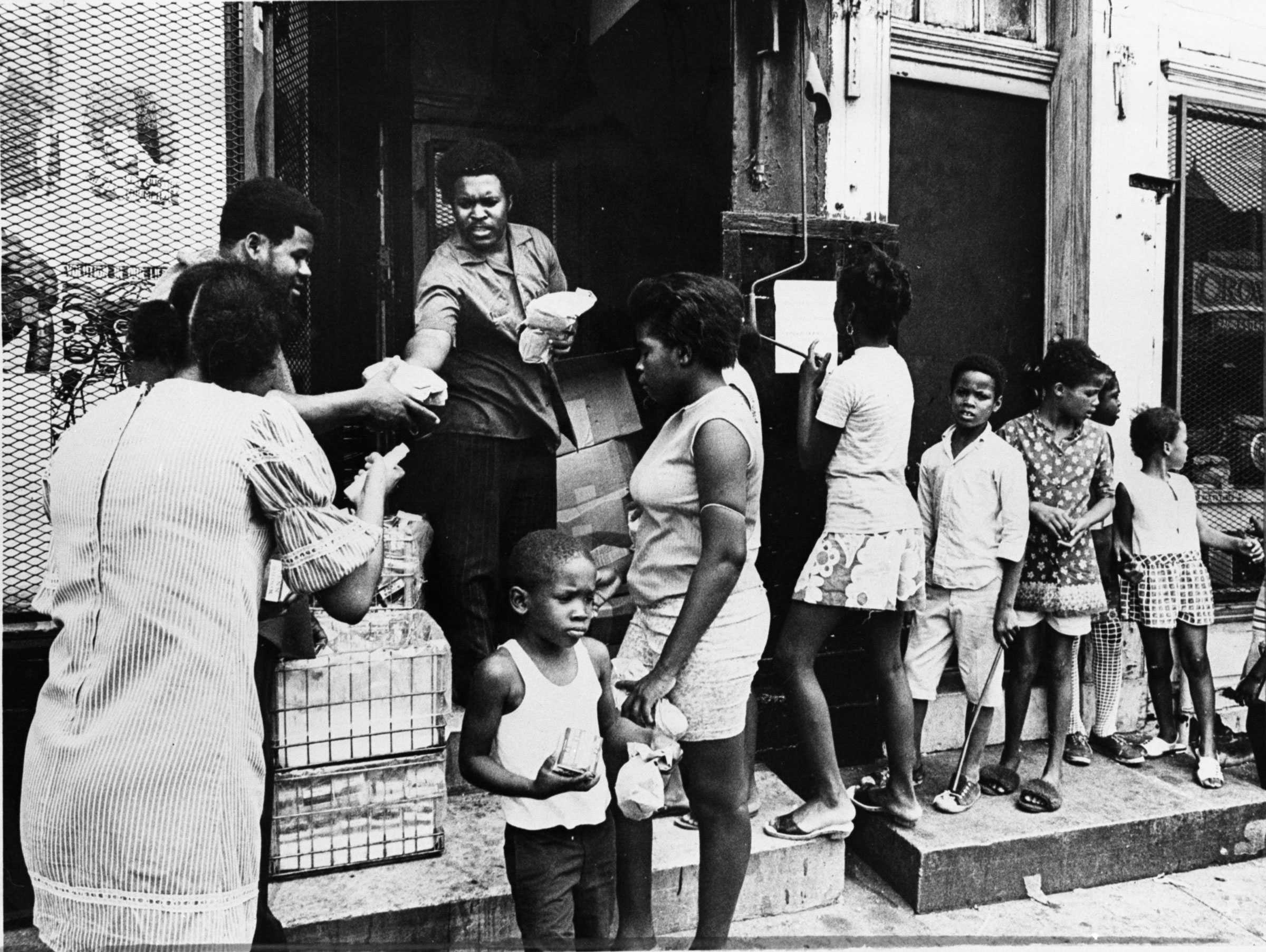 A member of the Black Panther Party distributing free lunches to North Philadelphia residents. A line of children line up against the building.