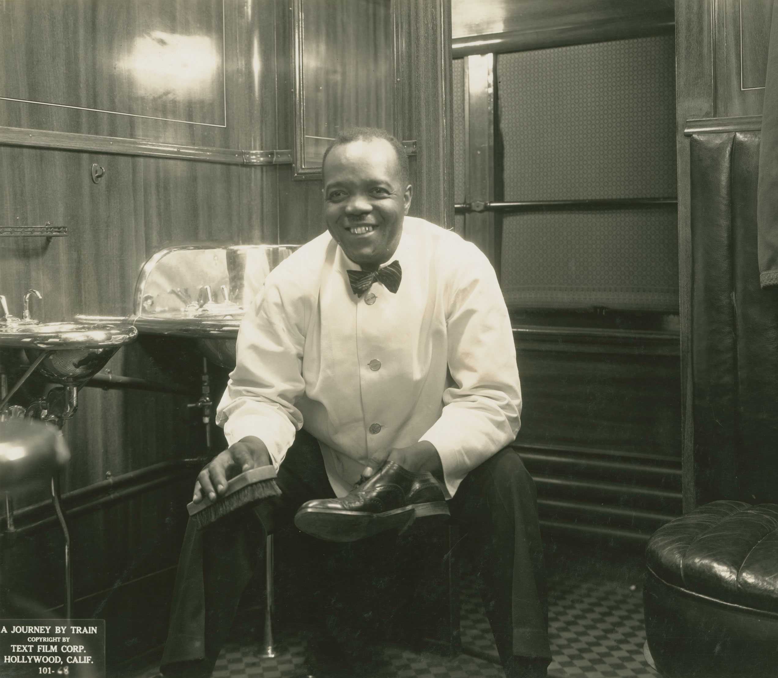 Silver gelatin print of a Pullman porter shining a shoe in a restroom train car, from the 1935 film A Journey by Train.