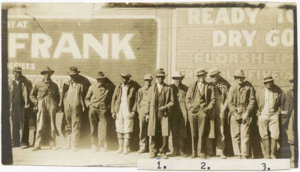 A photograph of a group of farmers stand side by side in front of a wall. The image is worn and slightly over exposed.