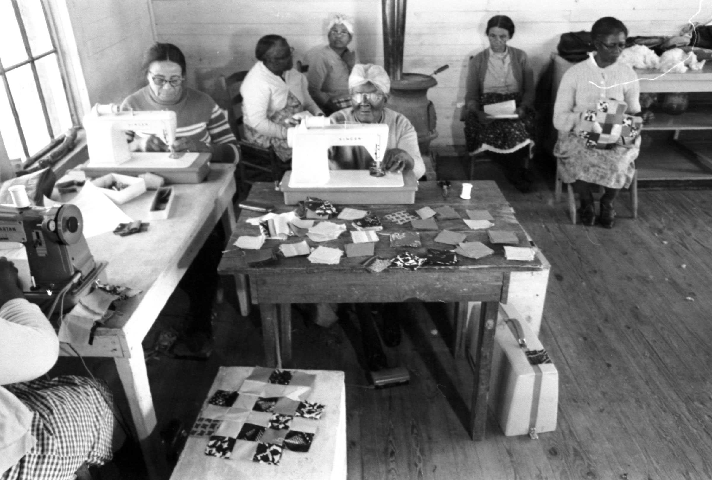 Black-and-white photo of women in a Poor People's Corporation sewing cooperative, making patchwork animals. Women sit at tables, sewing with machines, surrounded by fabric.