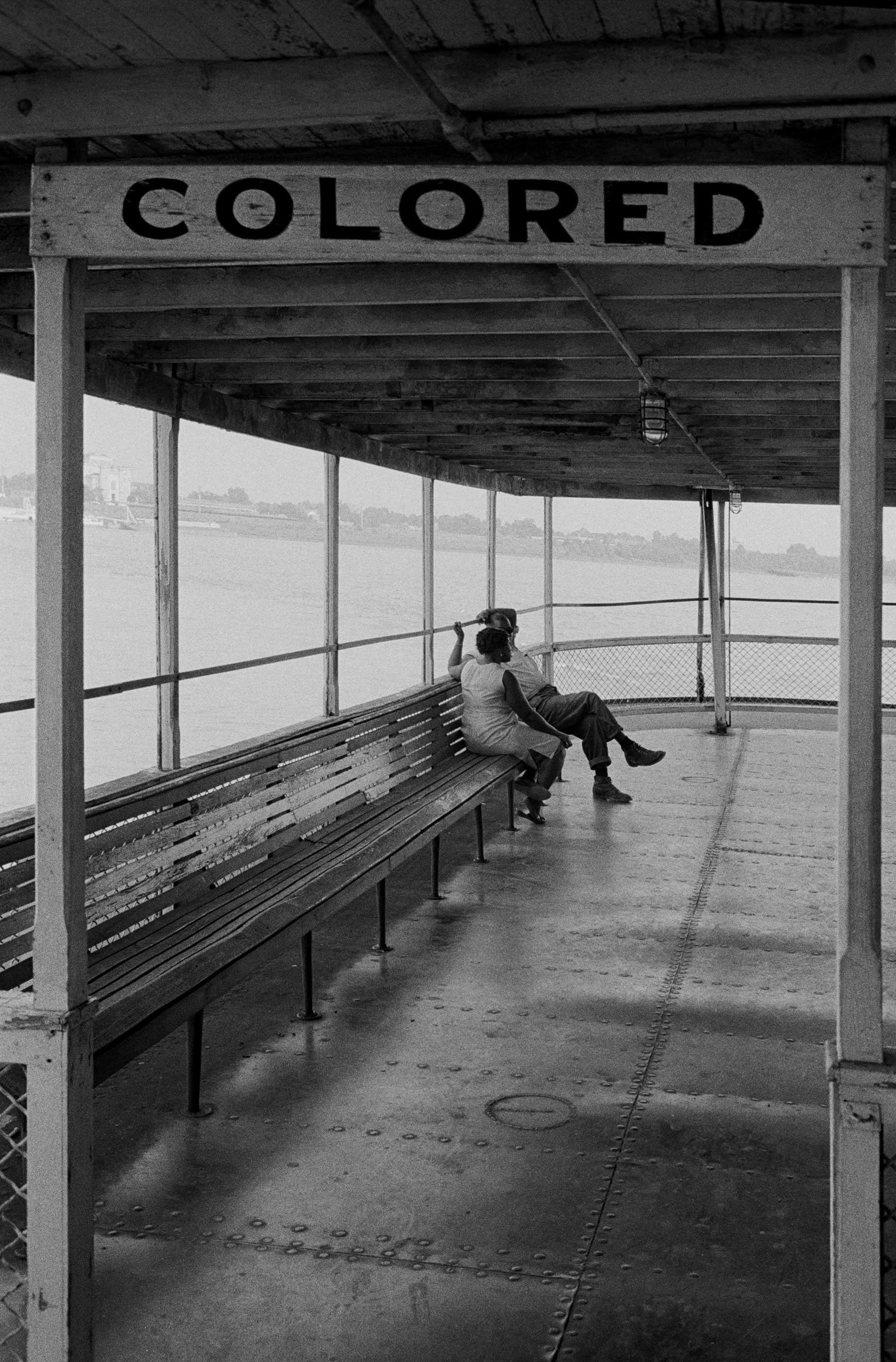 Black and white of a couple sitting on a bench in a waiting area marked "COLORED."  The waiting room appears to be for a boat/ferry as there is water visible in the background.