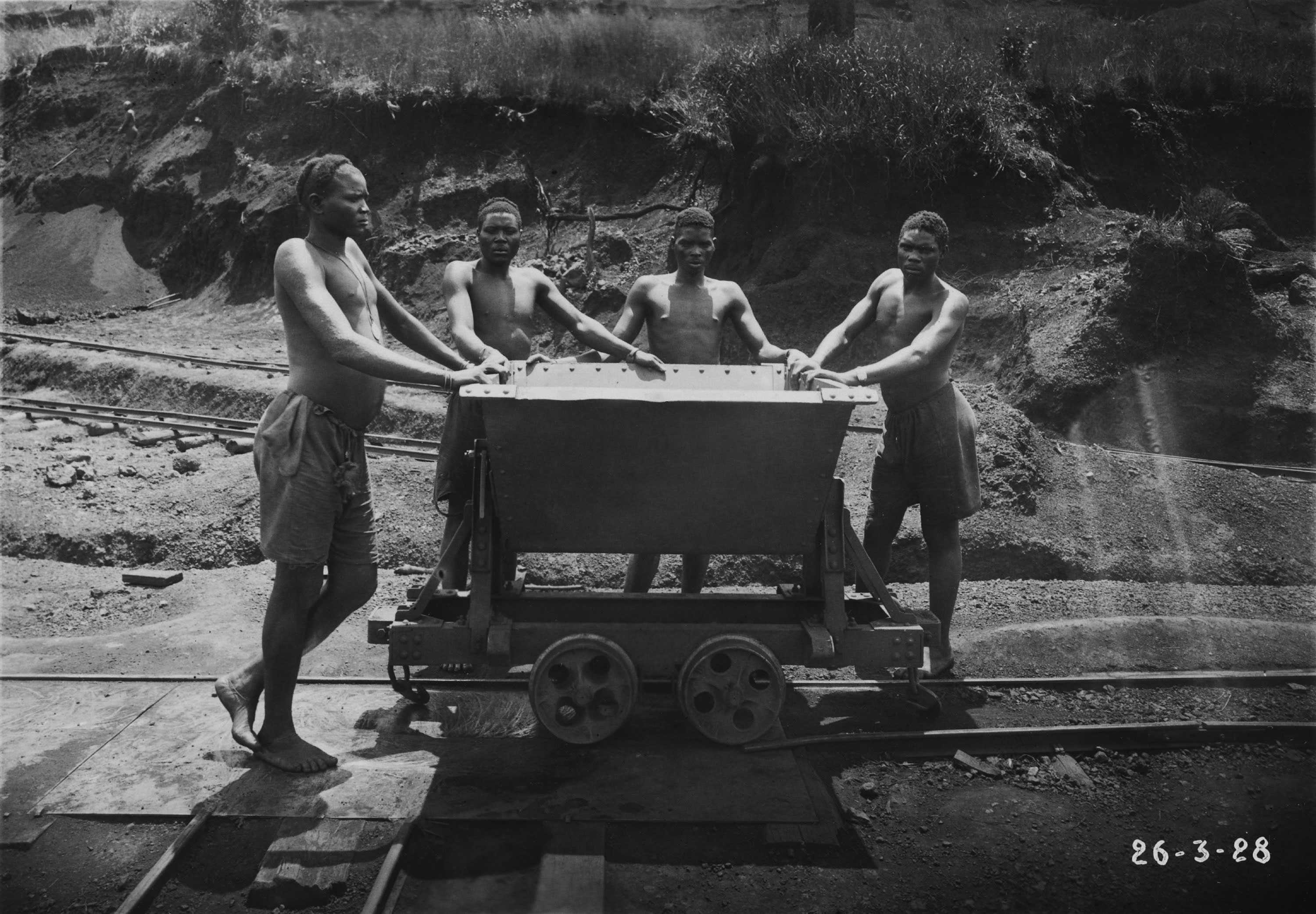 A black and white photo of 4 Ruandese workers push a mine cart on a track. The men are only wearing shorts without shoes.