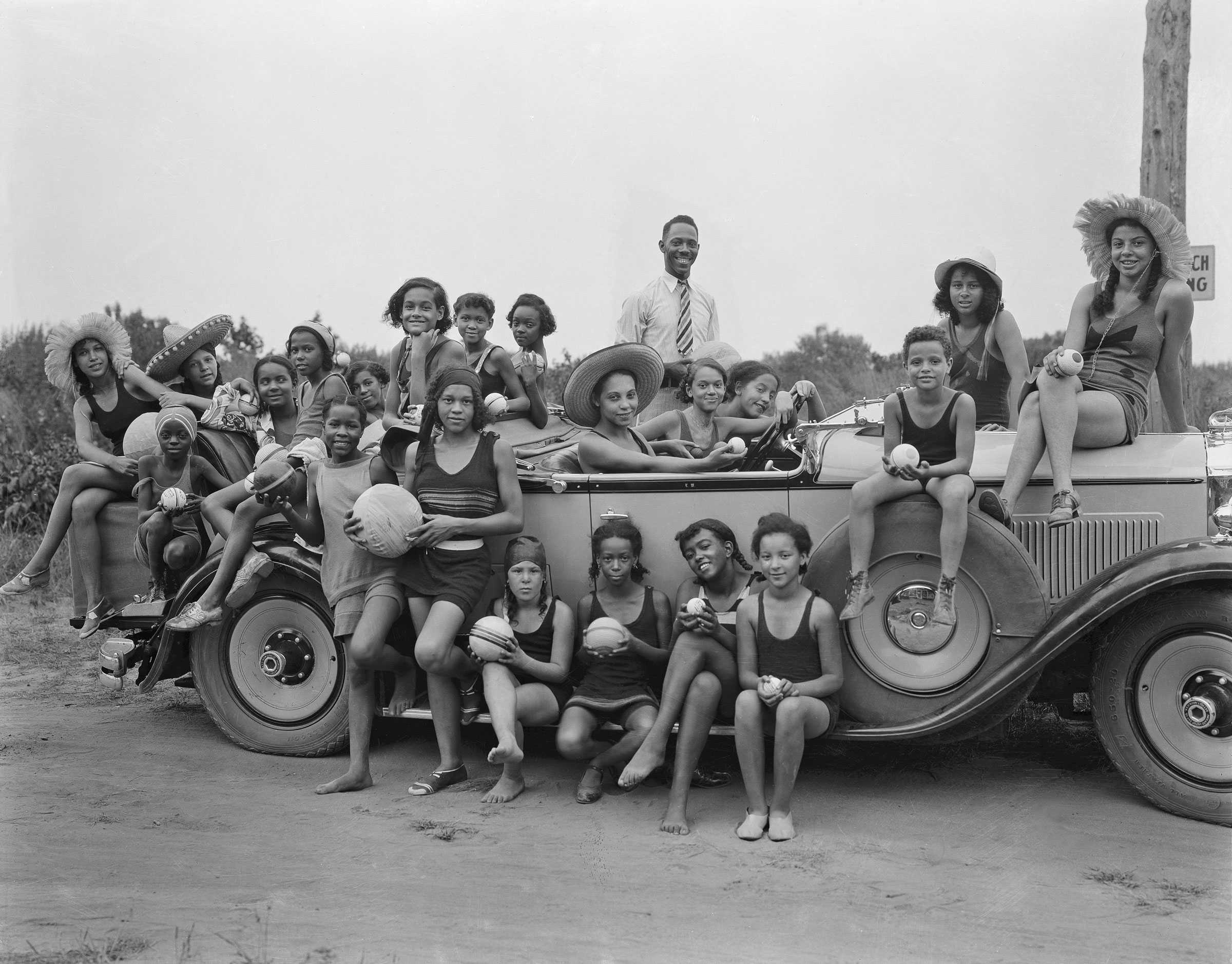 Black and white photograph of African American girls dressed in swimsuits pose in/on/around a car.  Some of them are holding balls and other sporting equipment.