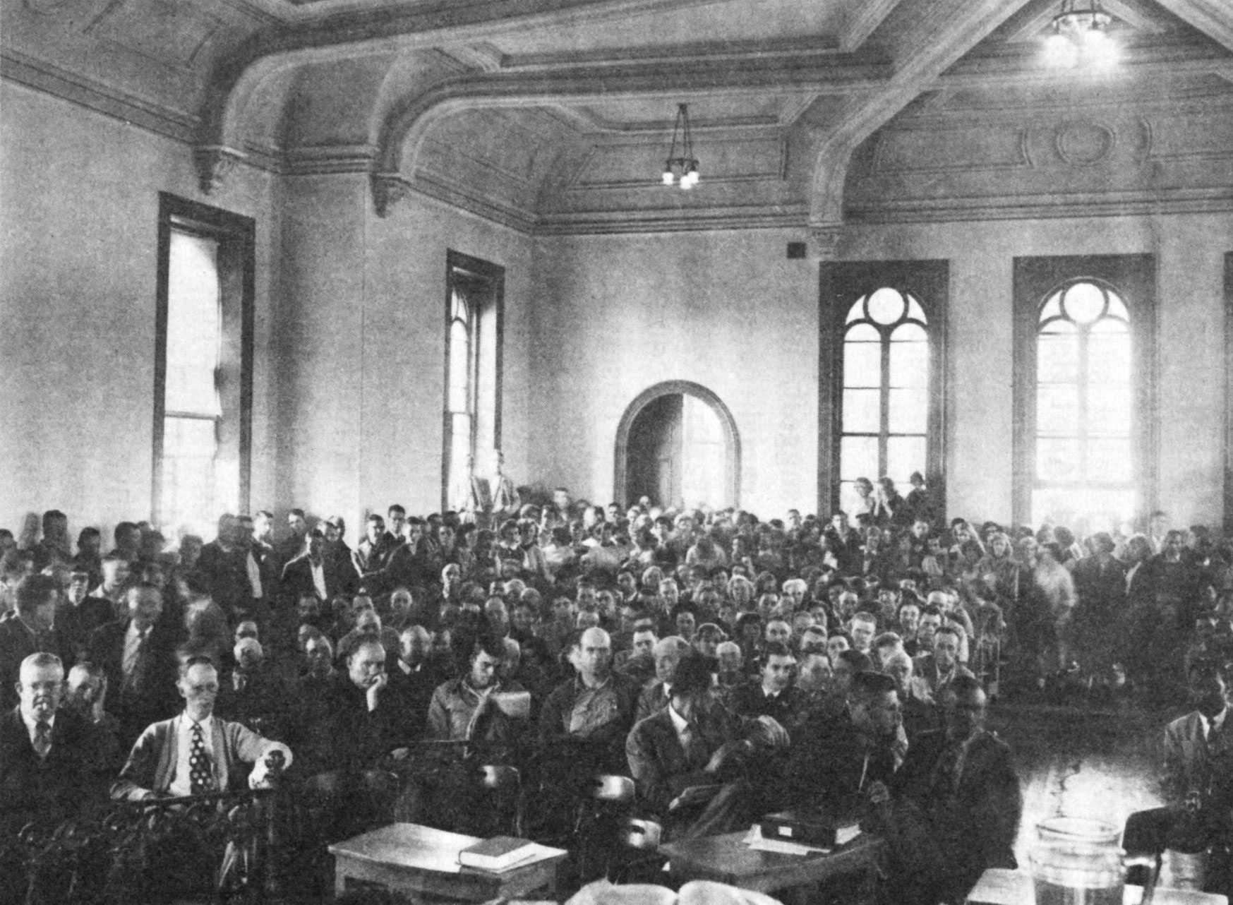 A black-and-white photo of a crowded courtroom watching Mack Ingram's trial. People are seated and standing, with arched windows and high ceilings in the background.