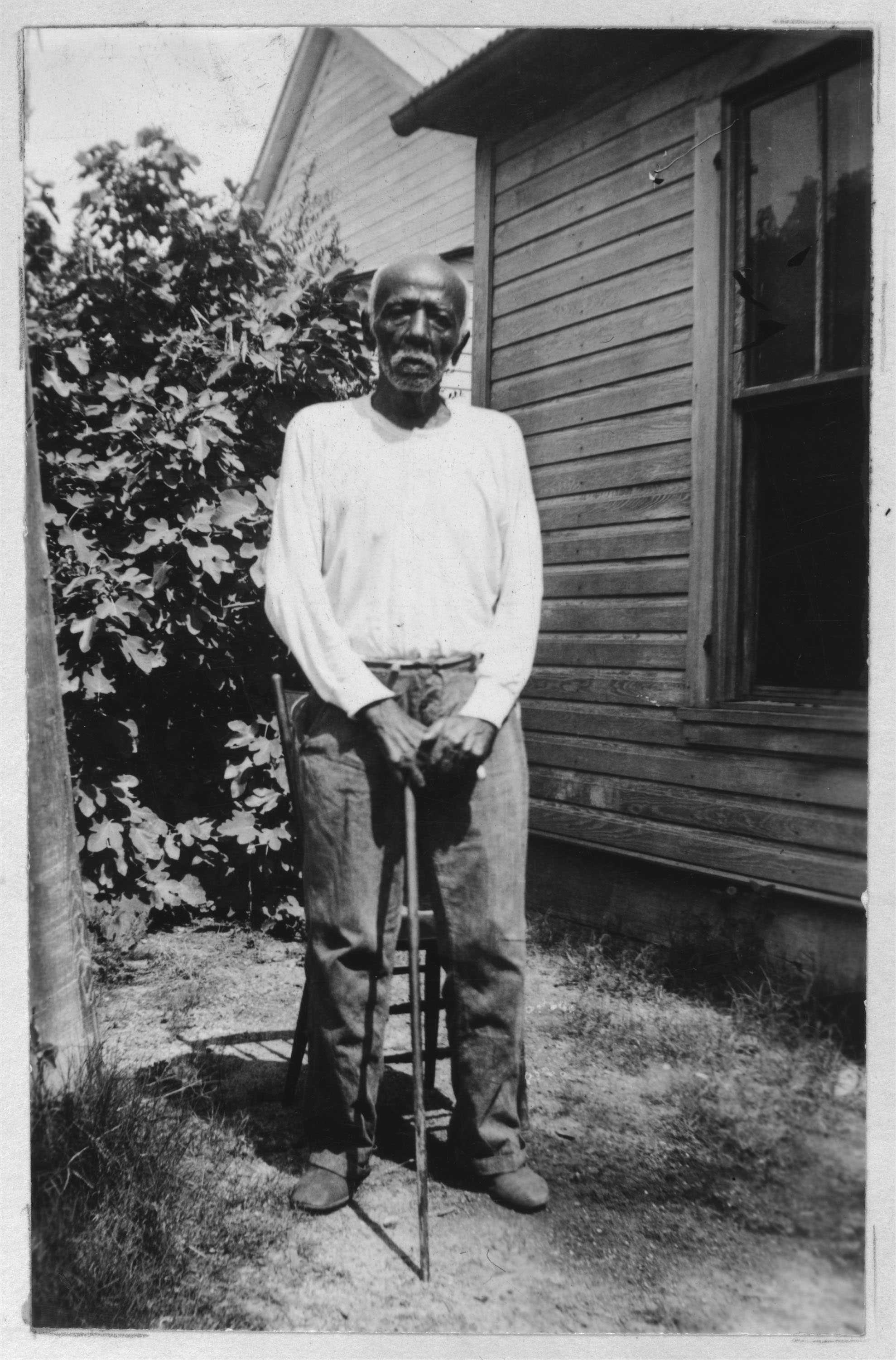 A black and white portrait of Martin Jackson, standing with a cane. He is wearing a white long sleeved shirt.