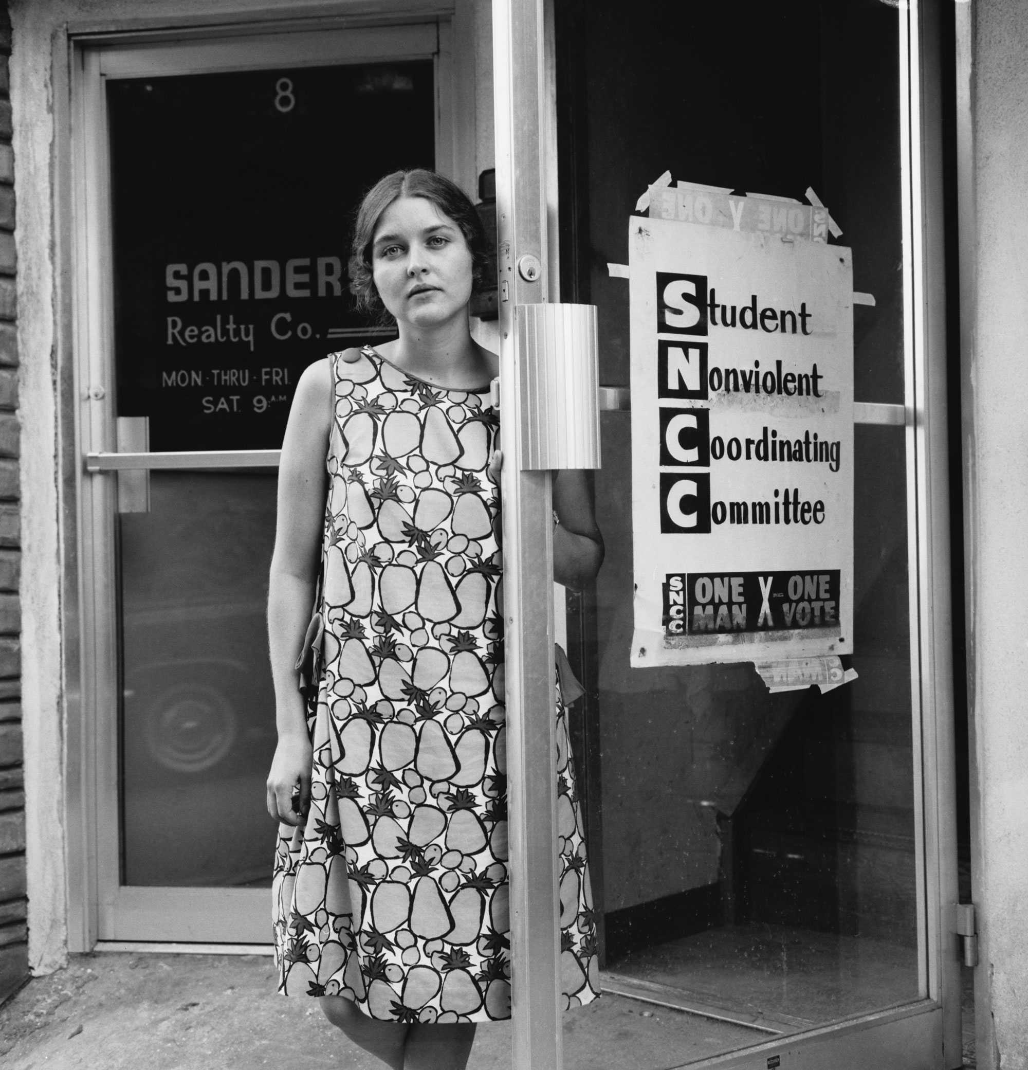 Constancia Romilly poses for a picture, holding the glass do leading to the SNCC headquarters. She is wearing a patterned dress with a pineapple graphic design.