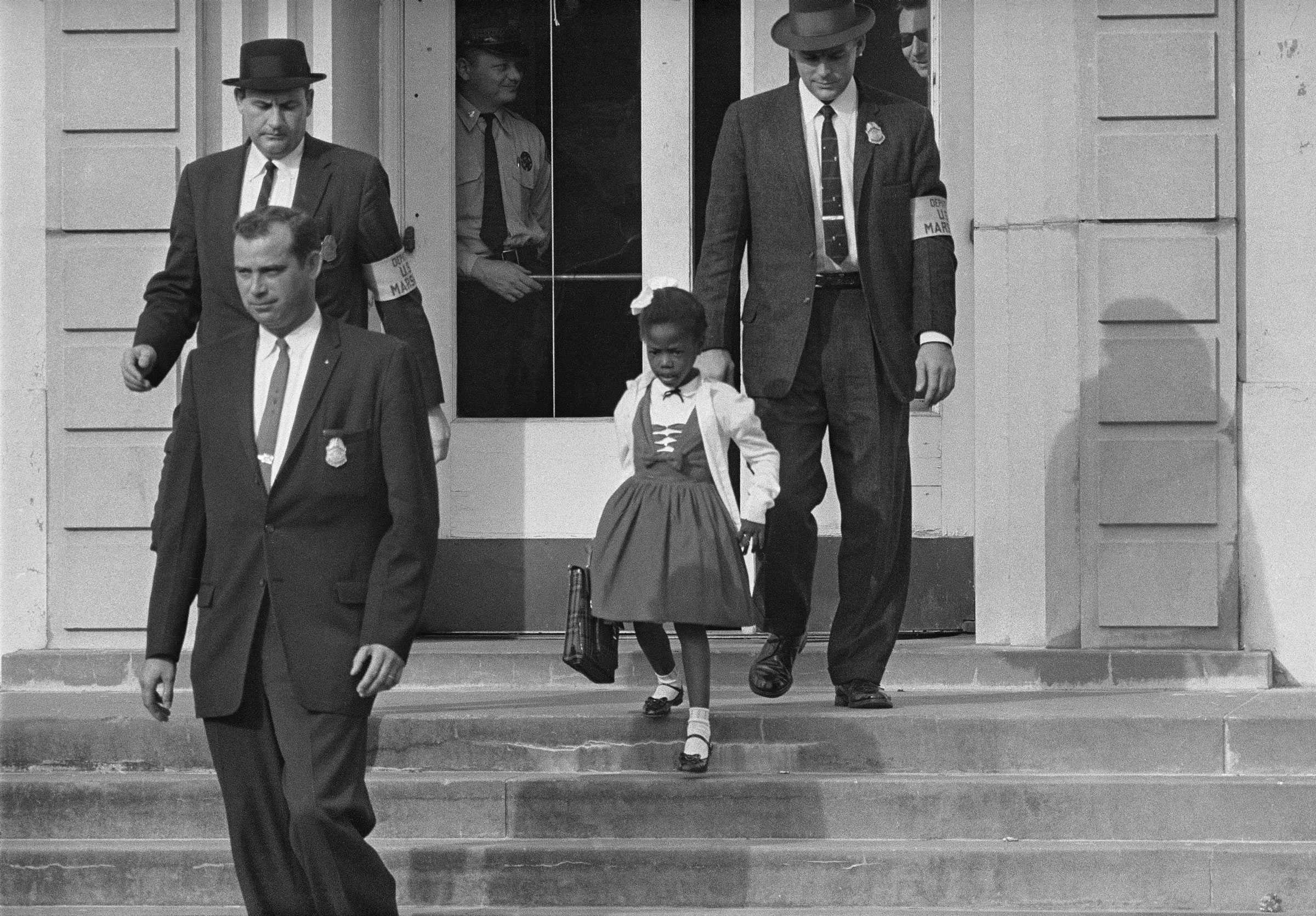 A black and white photograph of Ruby Bridges being escorted by three U.S. deputy marshals as she leaves school.