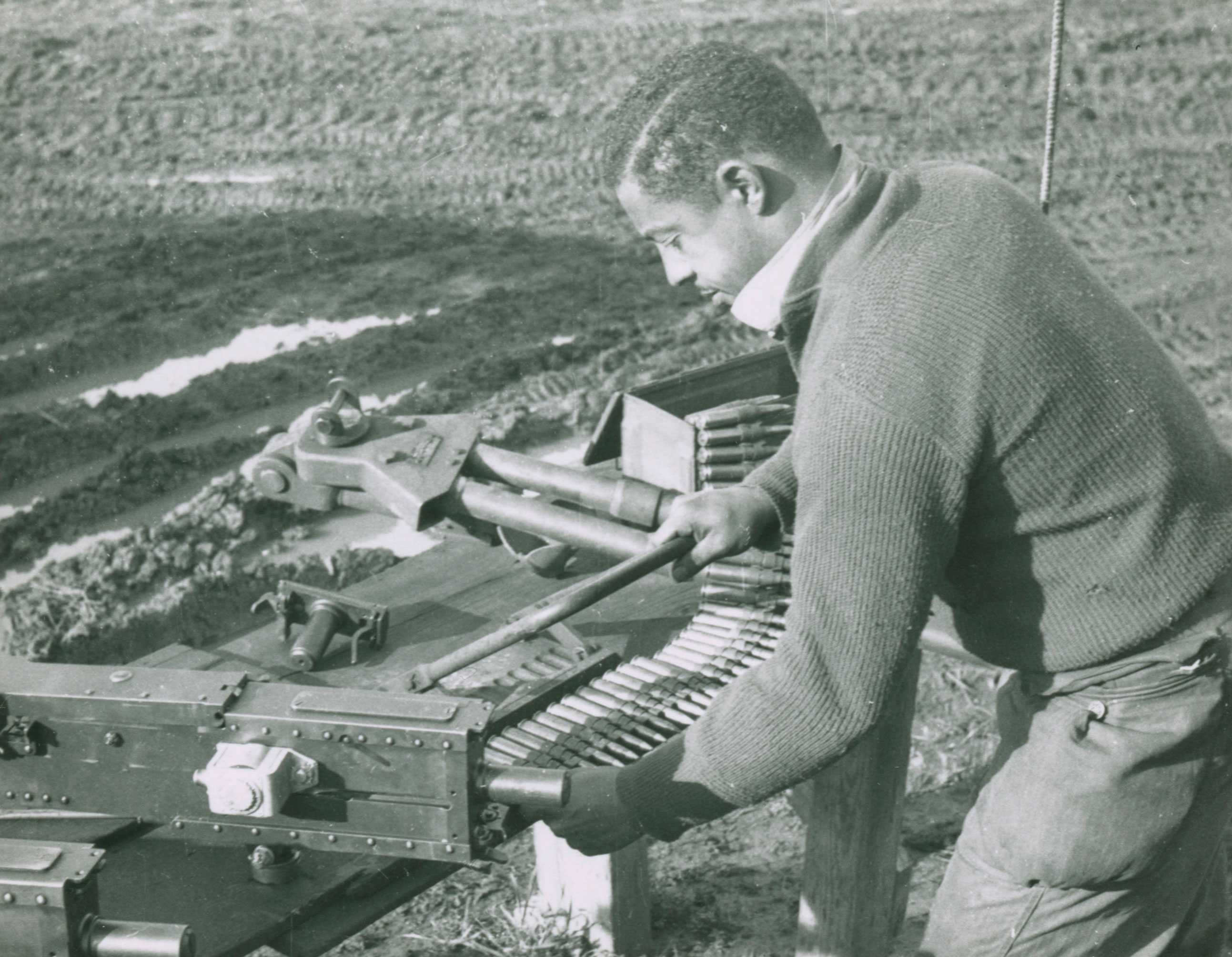 A black and white photograph of a Tuskegee airman working with tools and ammunition on a low wooden bench.