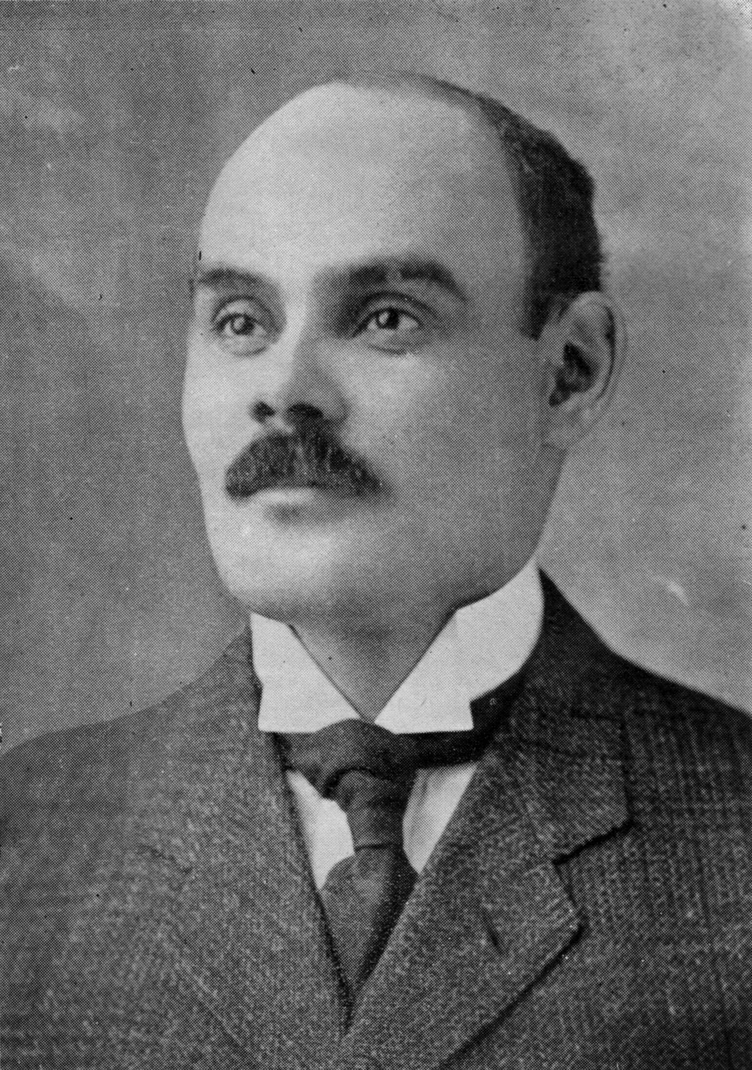 Black and White portrait of a balding man with a mustache.  He is dressed in a suit and tie.