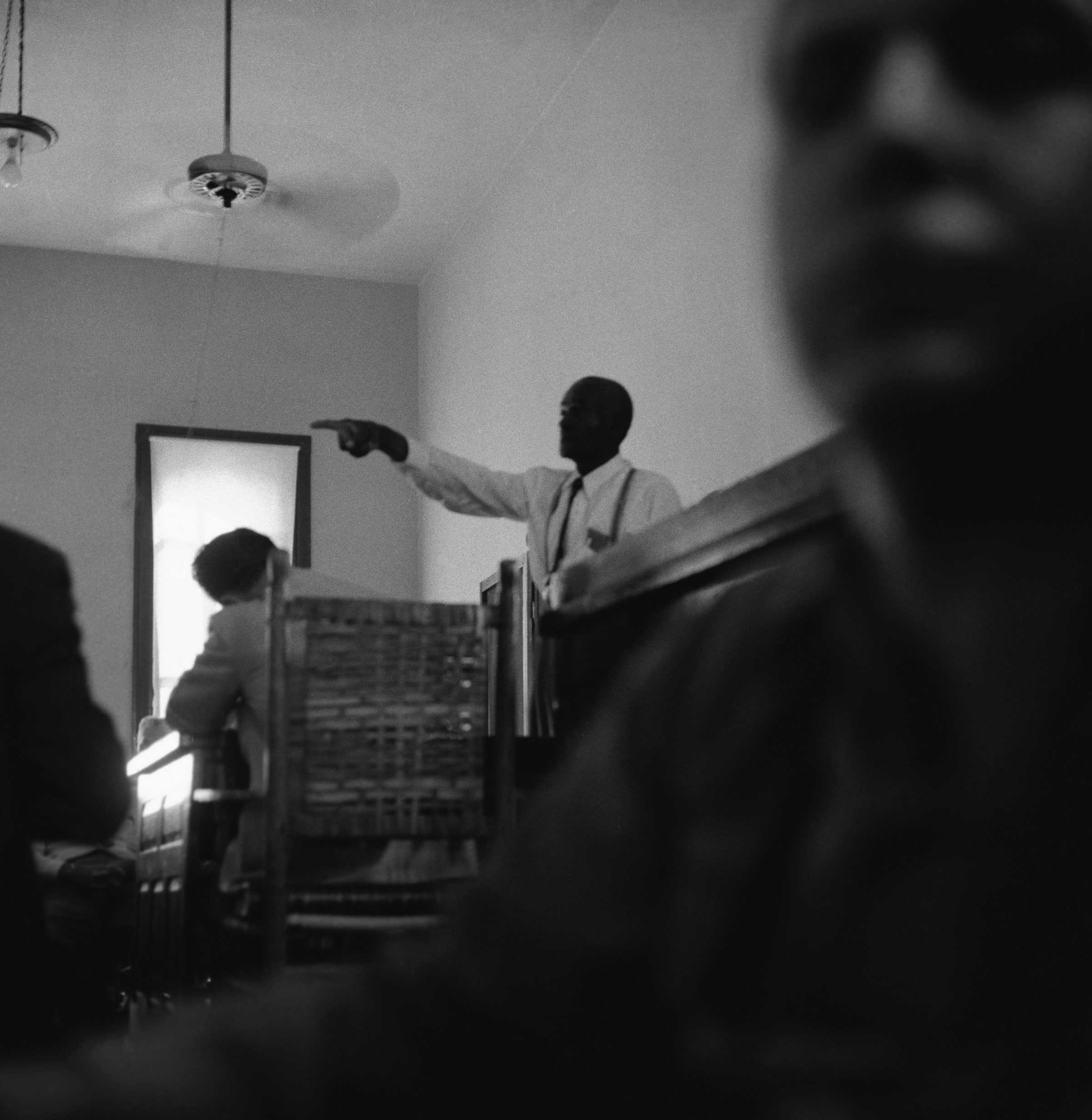 A black and white photograph of Mose Wright testifying on the stand in the background. A blurred figure in the foreground covers the right side.