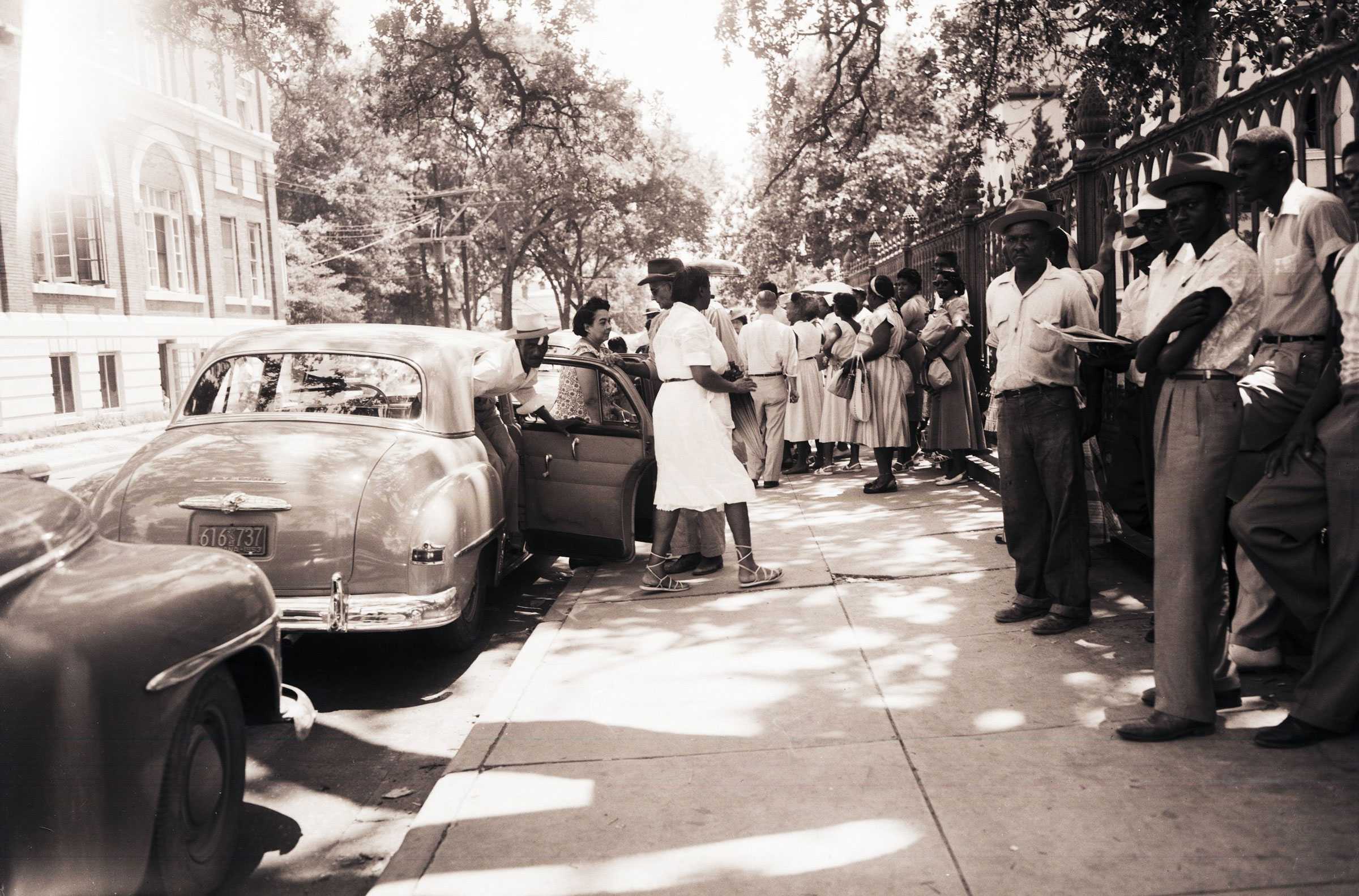 A black and white photograph of boycotters waiting on the streets to carpool. A group of people are getting out of a parked car.