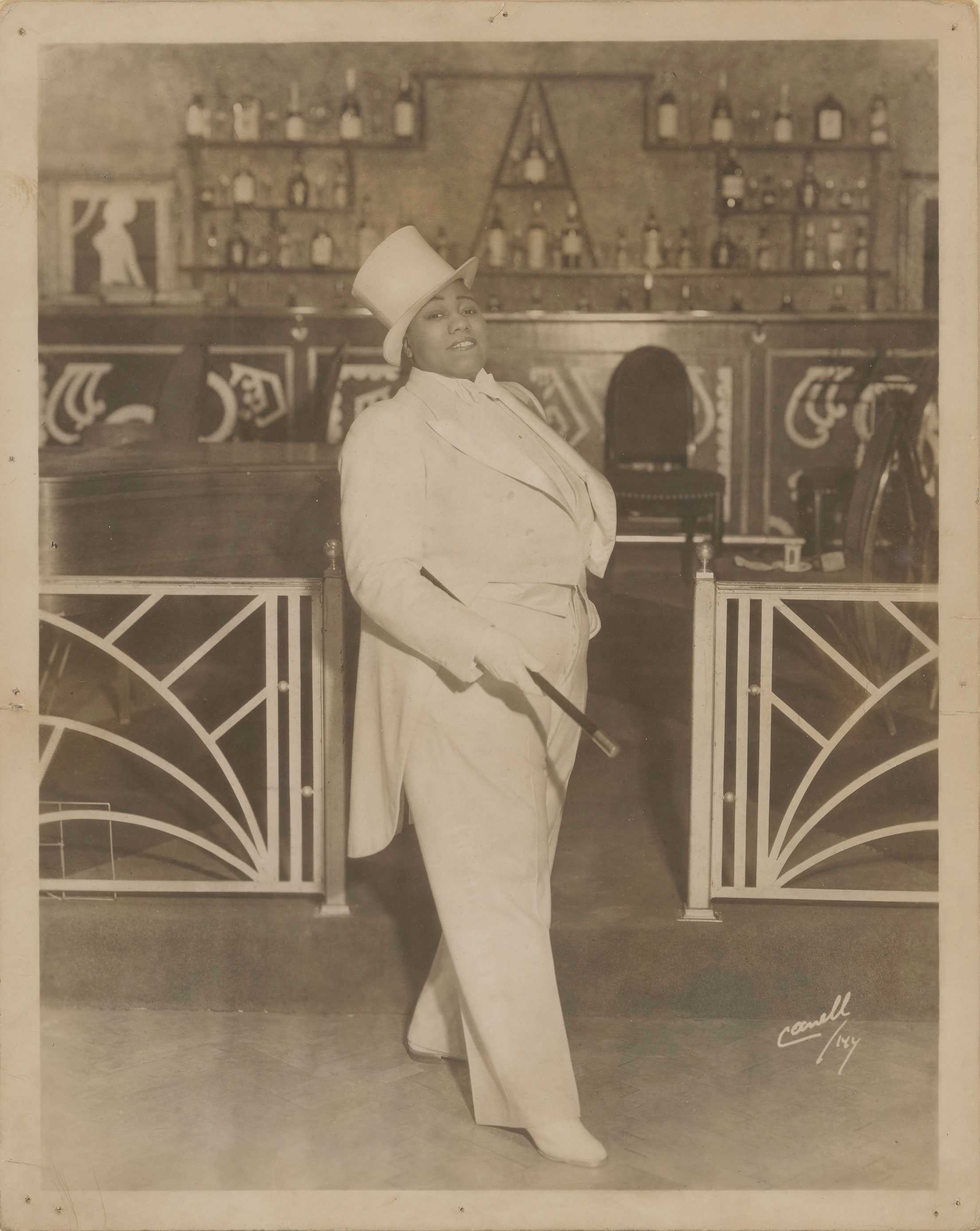 A sepia photography of Gladys Bentley in a white tuxedo and top hat, holding a cane in a bar.