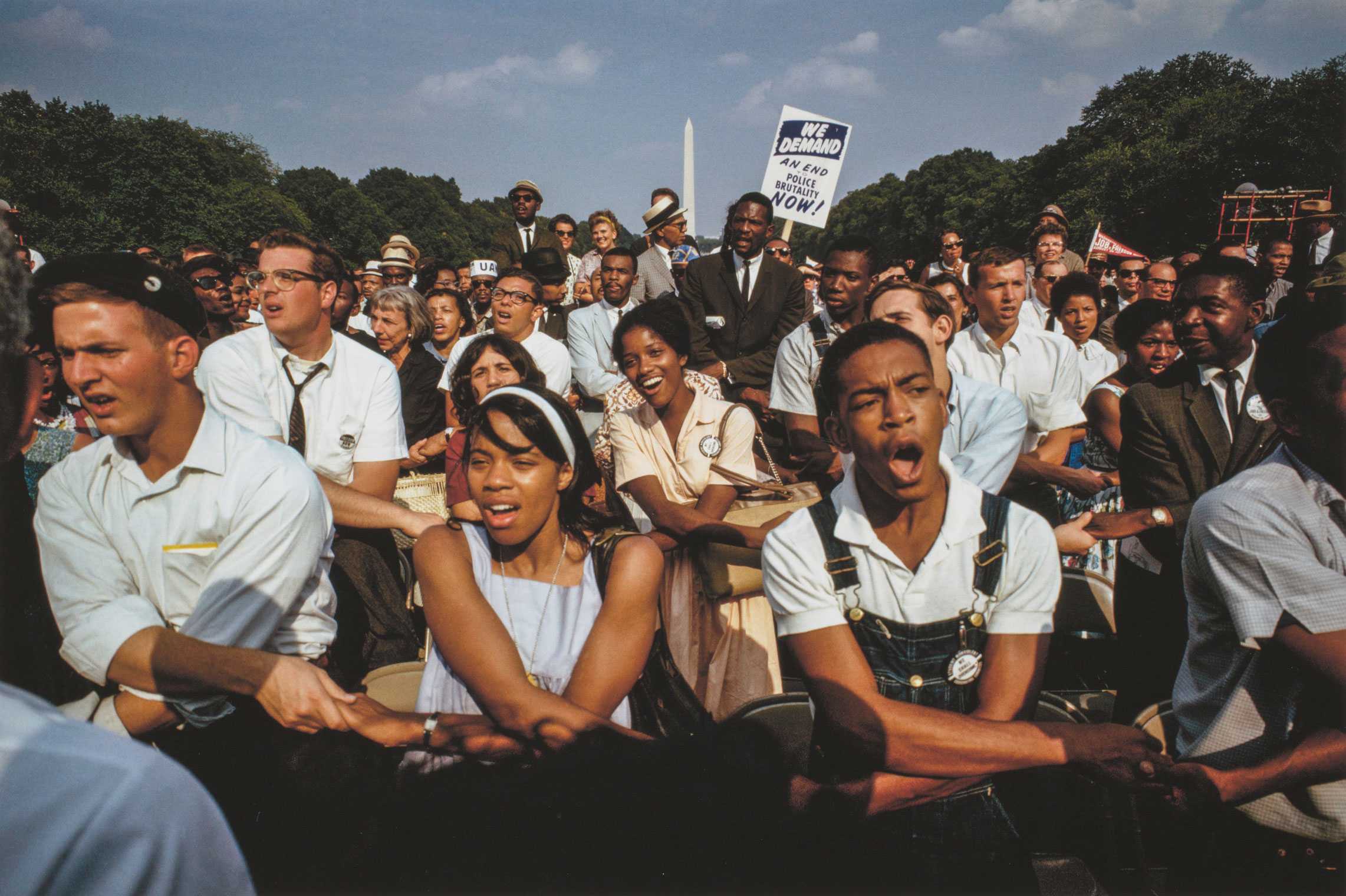 A color photograph by James P. Blair of the crowd of participants at the 1963 March on Washington. The image depicts a large crowd of different ages, genders and races gathered between the steps of the Lincoln Memorial and the Lincoln Memorial Reflecting Pool. The Washington Monument is visible in the far background. In the foreground of the image are rows of seated men and women with arms crossed in front of them and holding hands with the people on either side of them. Someone in the crowd holds up a placard that reads “WE DEMAND / AN END / TO / POLICE / BRUTALITY / NOW!” The print is signed by the photographer beneath the image at bottom right: [James P. Blair / 9 or 9]. Printed along the bottom edge is the caption and a copyright notice in black text.