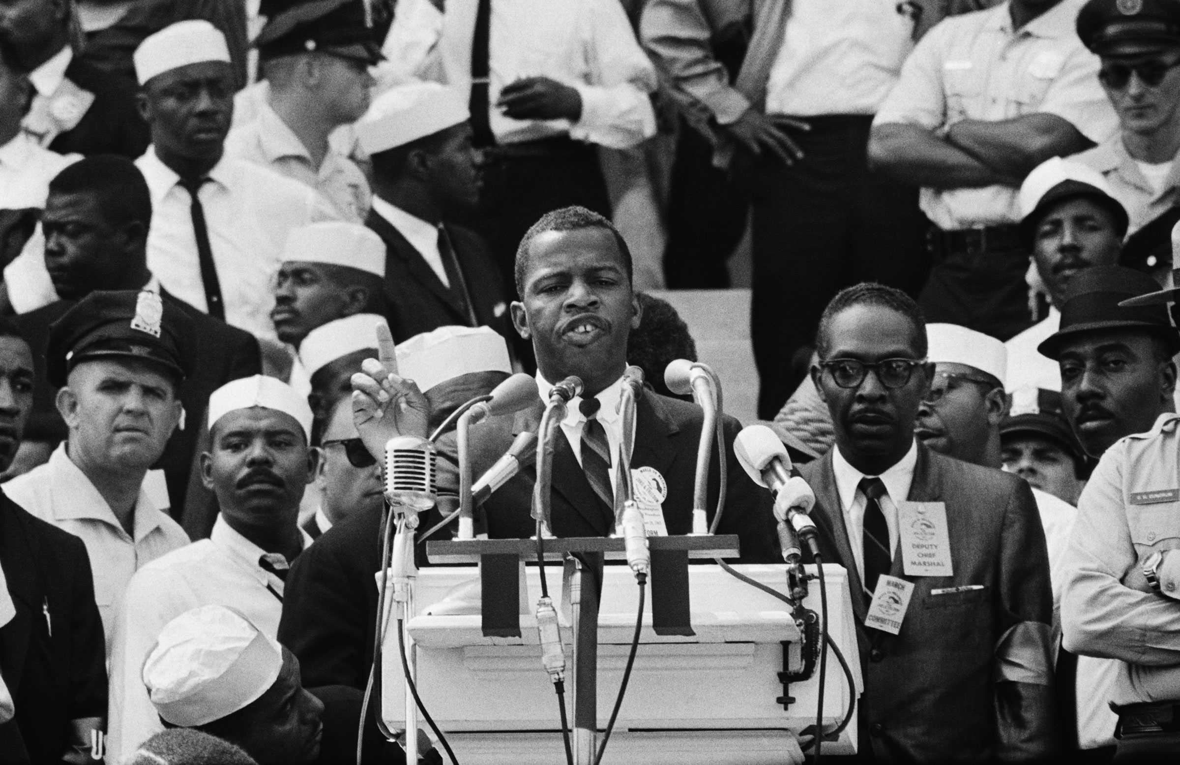 John Lewis speaking  into multiple microphones in front of a crowd at the Lincoln Memorial. Black and white civilians and police officers stand behind him on the stairs.