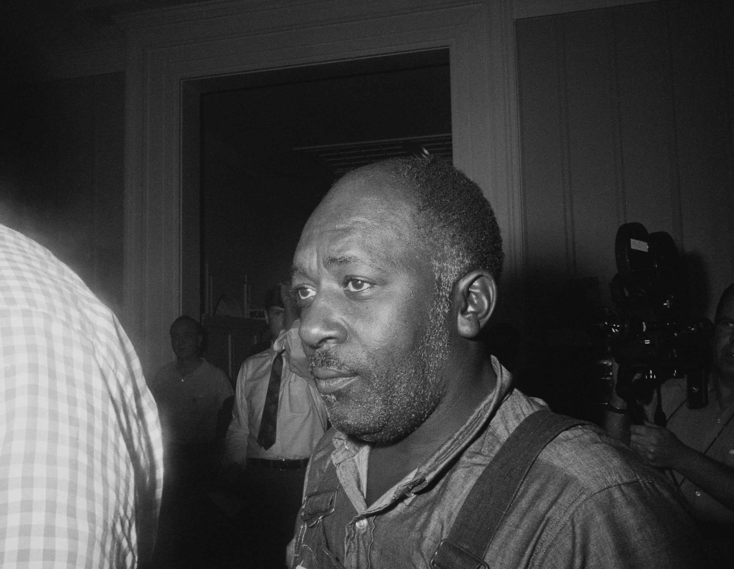 A close photograph of Charles Sims walking through a crowd at City Hall. He is dressed in overalls, while the others are dressed in dress shirts.