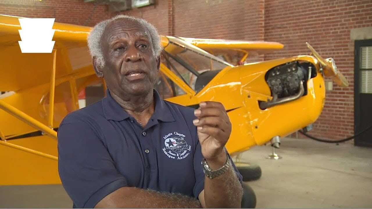 An older man sits for an interview in front of a bright yellow airplane