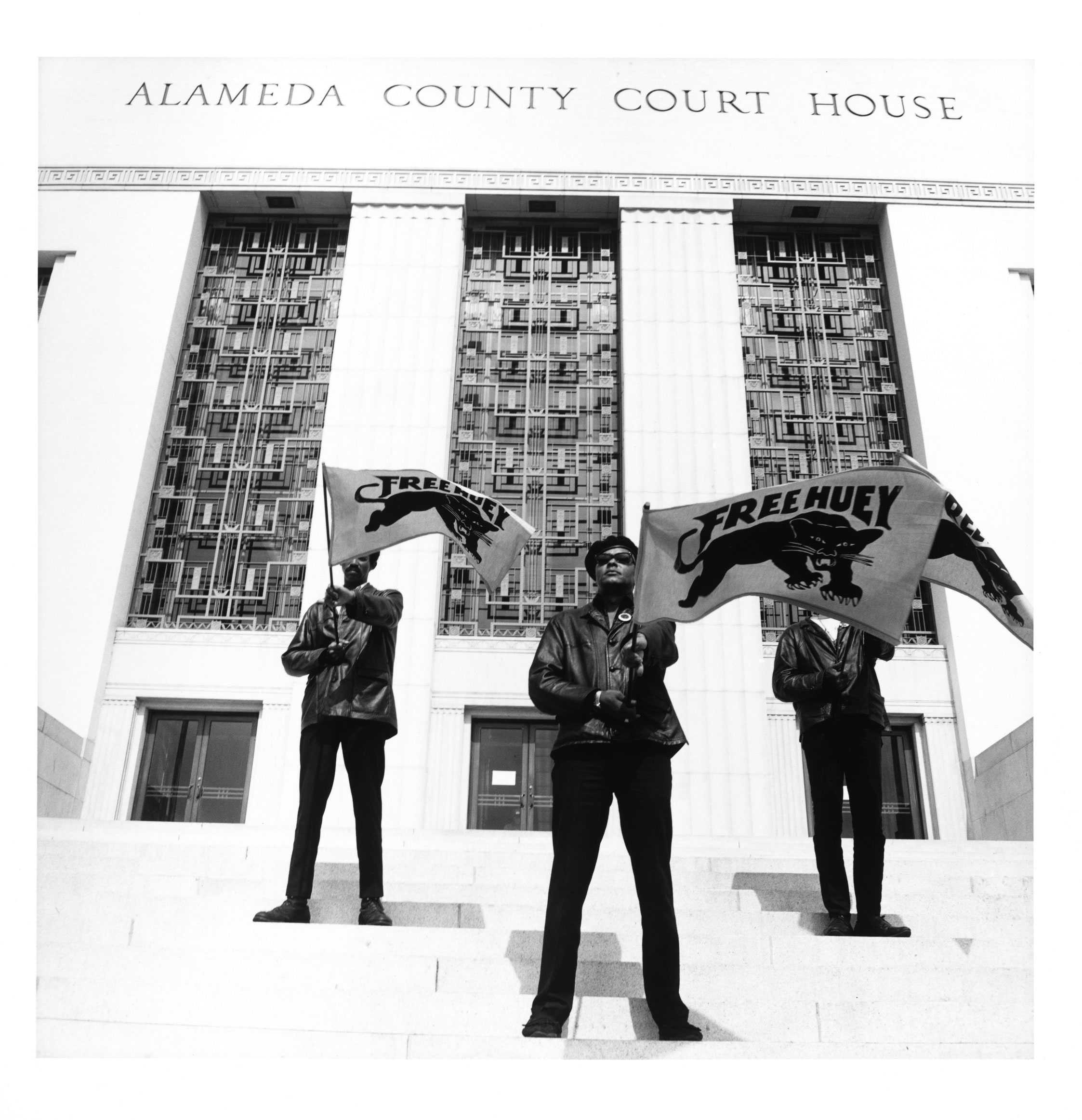 A black-and-white image of 3 African American men wearing leather jackets and berets and carrying Free Huey banners on the court house steps.