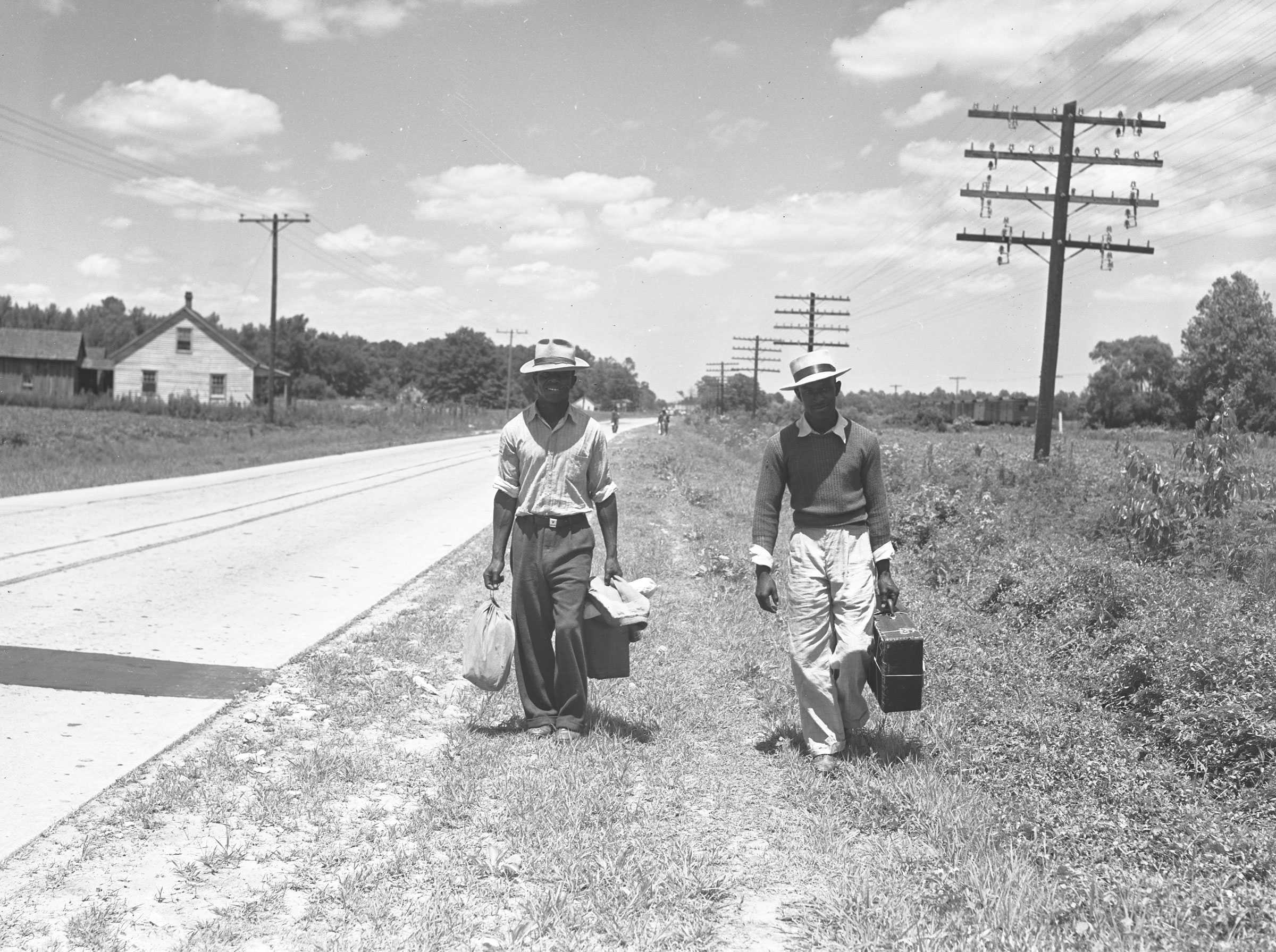 Black and white photograph of two men walking with suitcases along side of a road.