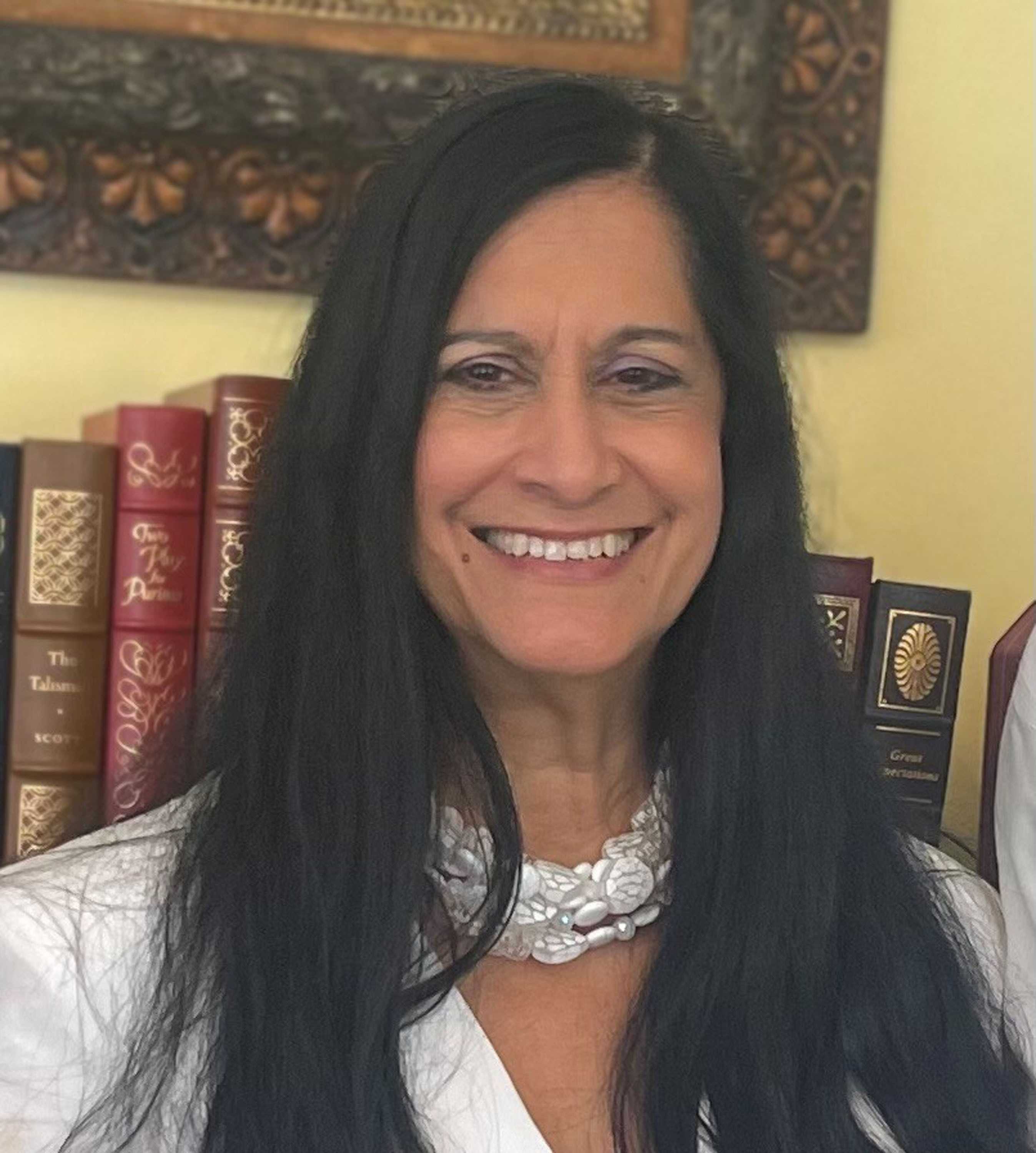 Color portrait photograph of  smiling woman with long dark hair.  She is wearing a white shell necklace and a white shirt.