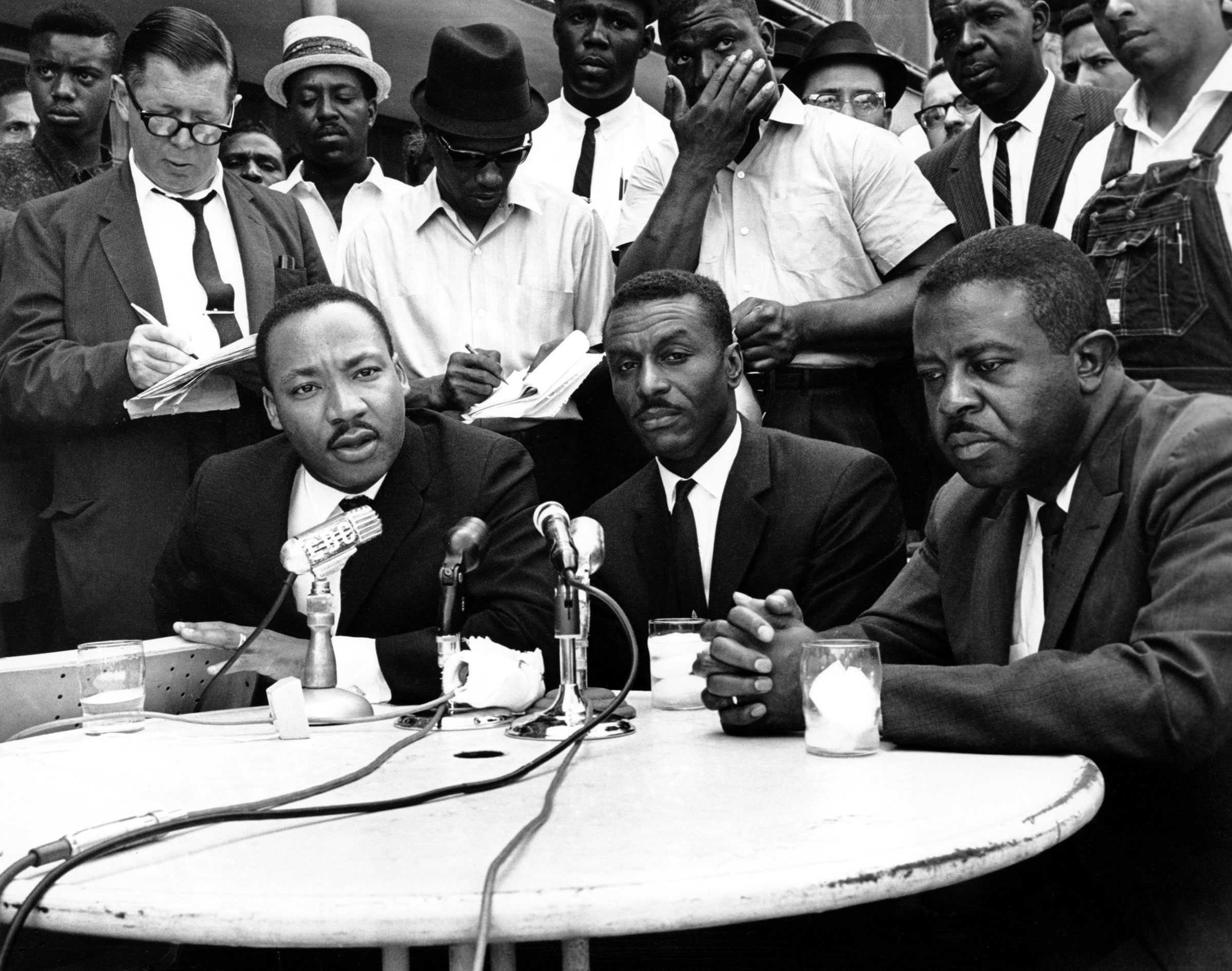 Martin Luther King Jr., Fred Shuttlesworth, and Ralph Abernathy speak into microphones at a press conference. A crowd of press stands closely behind them.