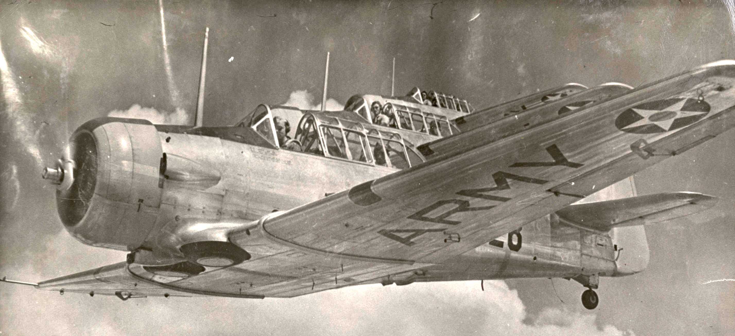 Three AT-6 training aircraft fly in formation at Tuskegee Army Air Field, Alabama, with pilots looking at the camera.