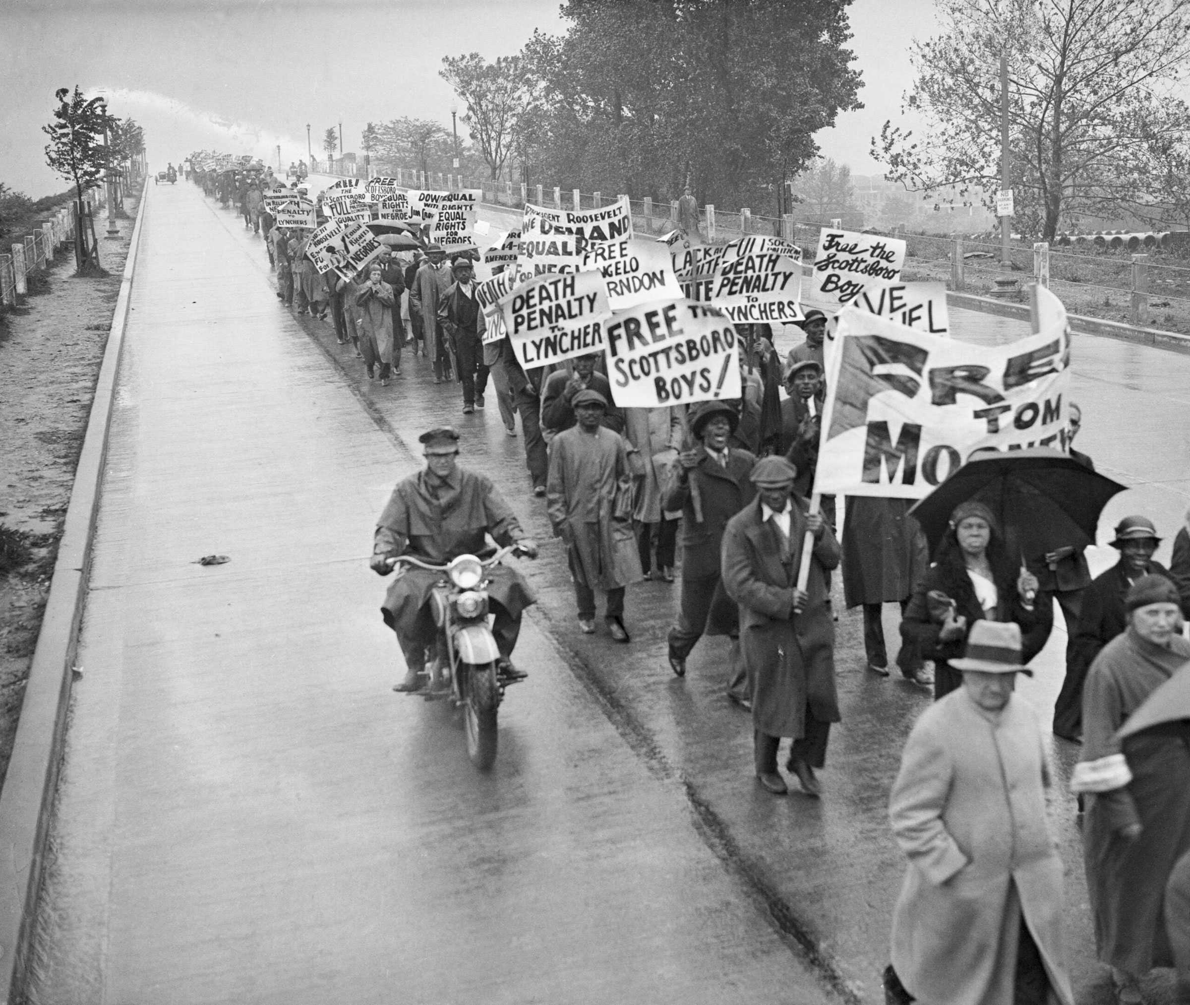 Black and white photograph showing a protest demonstration.  Marchers form a long line and hold signs "FREE THE SCOTTSBORO BOYS" and "DEATH PENALTY TO LYNCHERS."