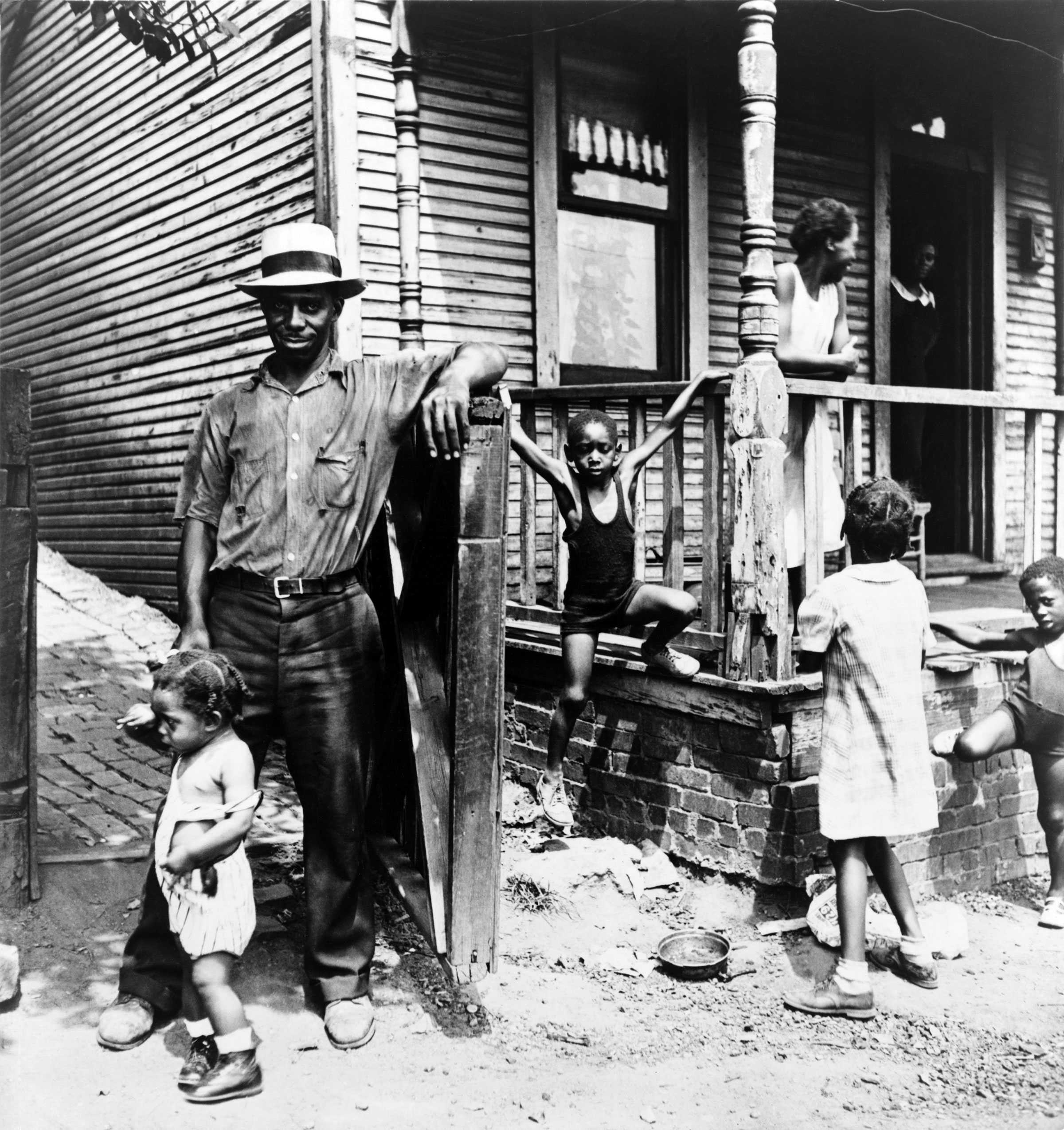 Black and white photograph of a Black family pictured outside their home.  There is a woman visible in the doorway, another one on the porch and four children