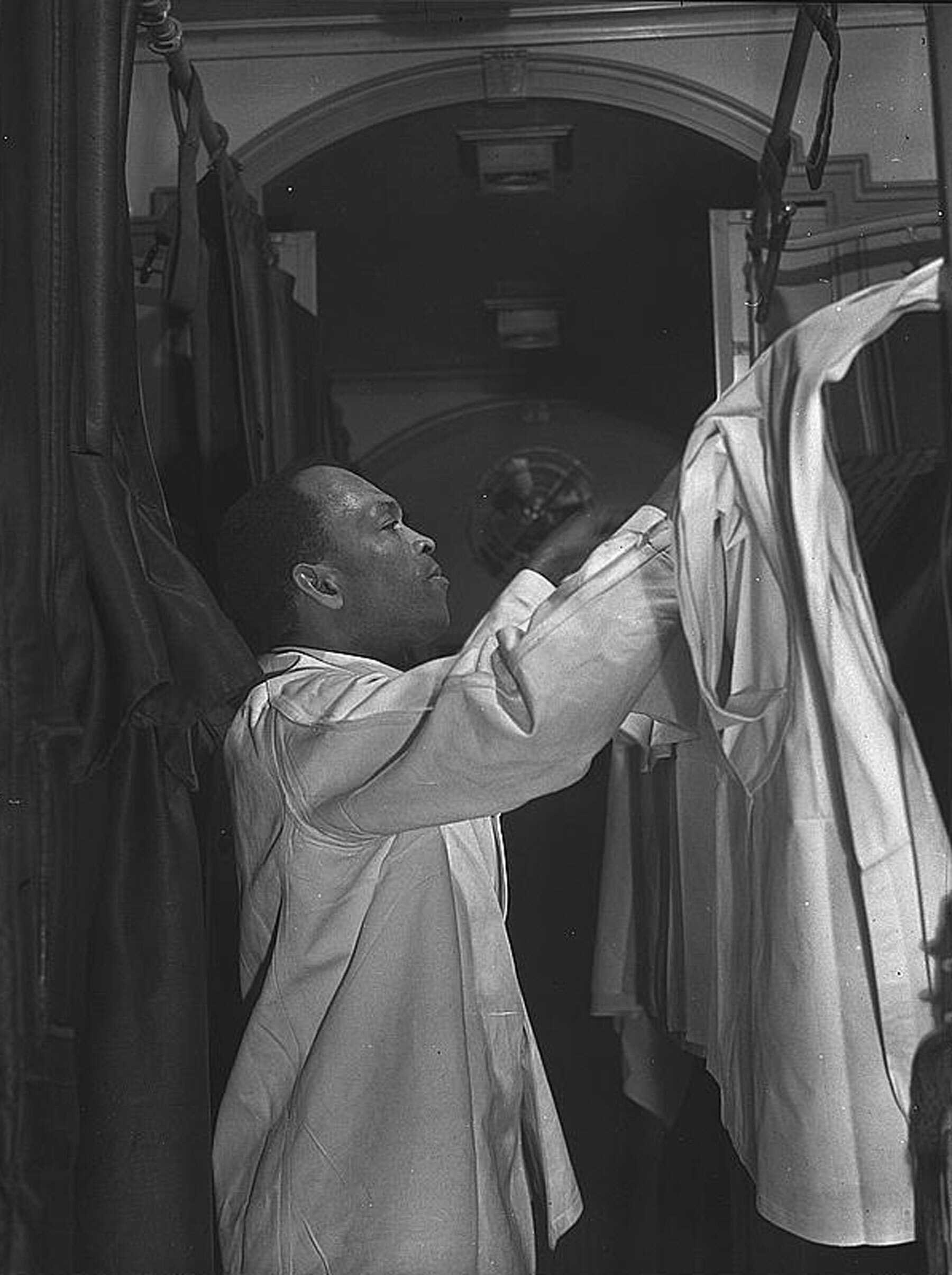 Black and white photograph of man making the bed of an upper bunk in a train compartment.