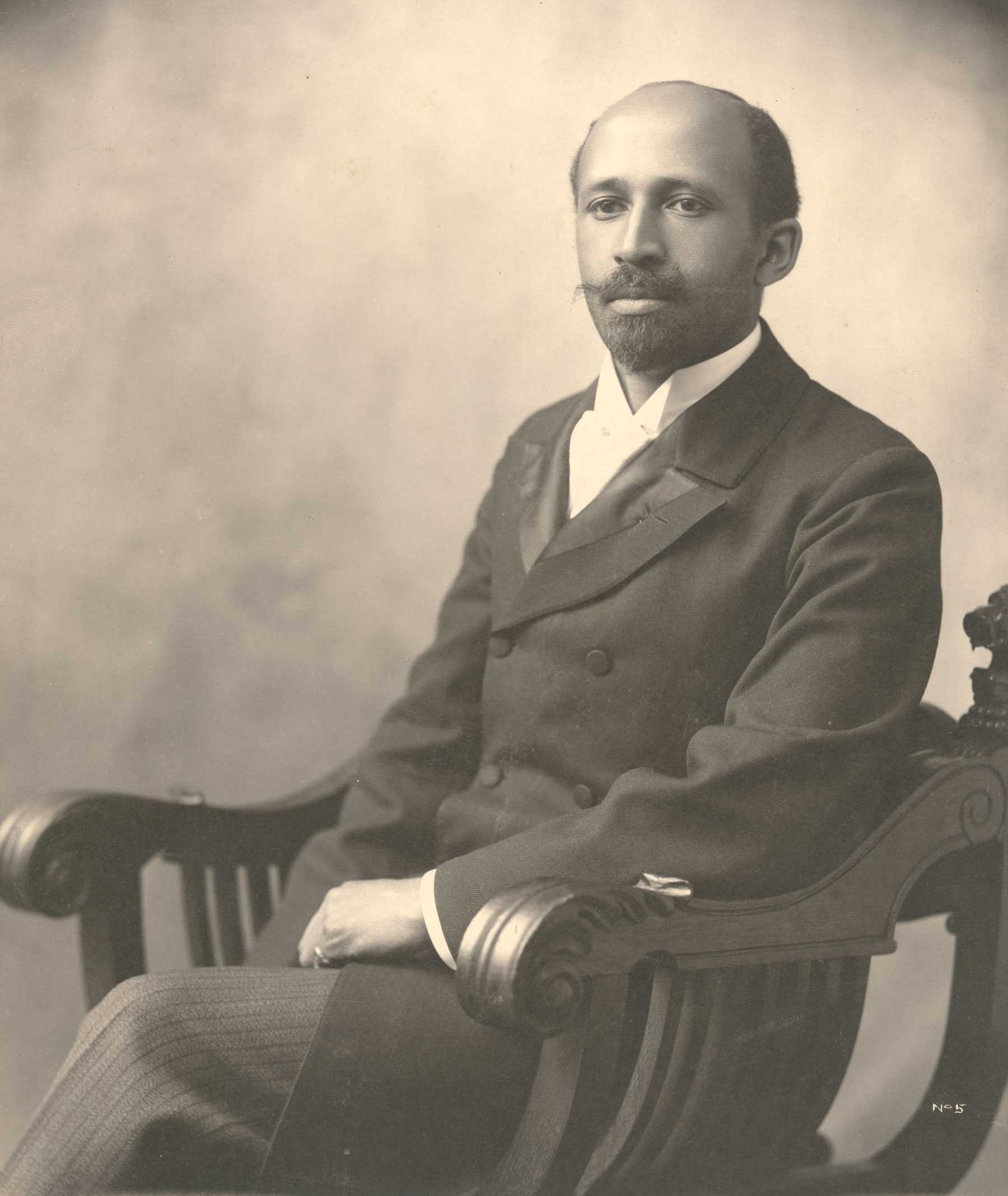 Black and white portrait of an African American male seated on an ornate wooden chair. The man is wearing a formal suit with a tie, and their hands rest on the chair’s arms.