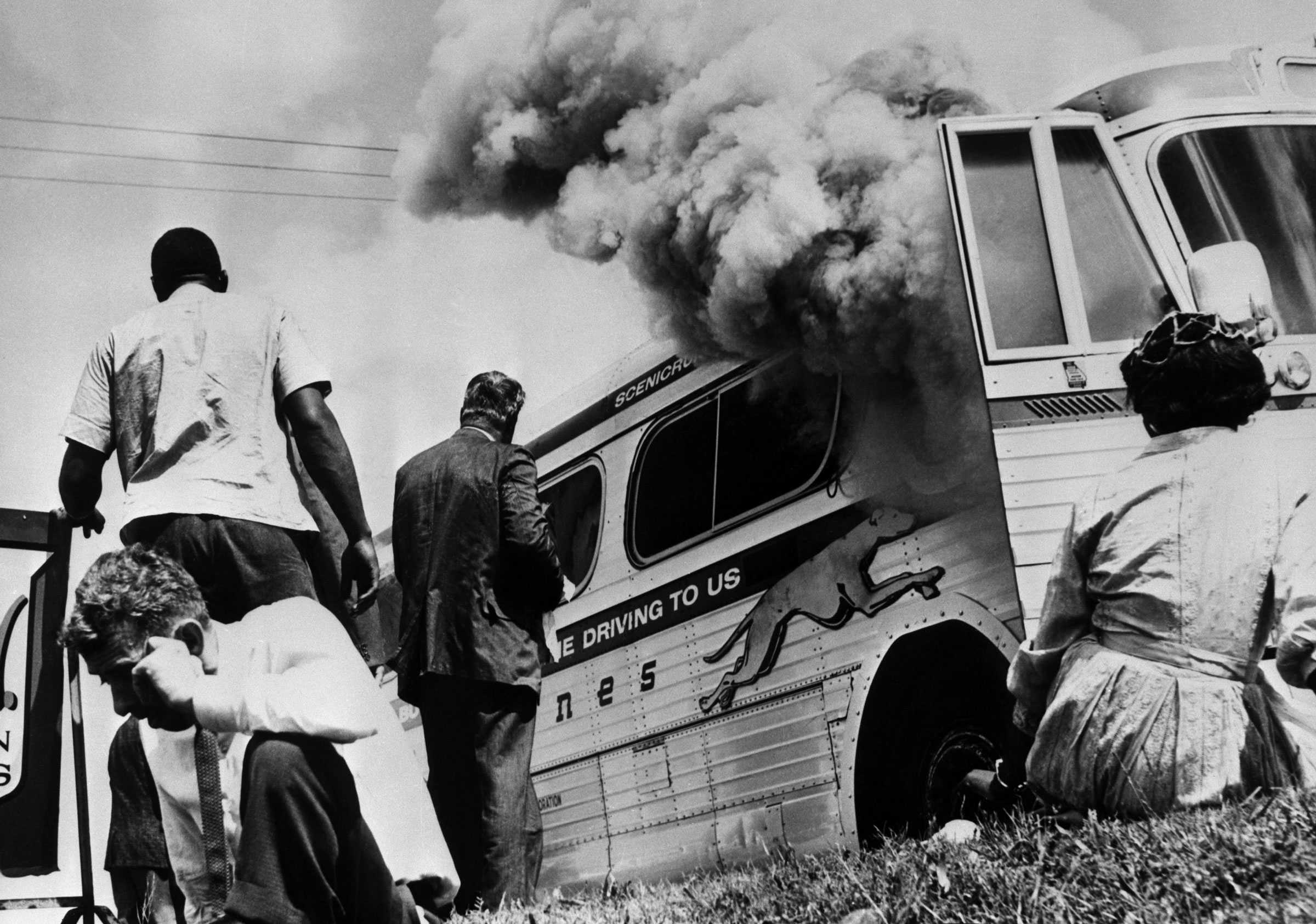 A black and white photograph of Freedom Riders sitting on the grass outside of their burning bus.