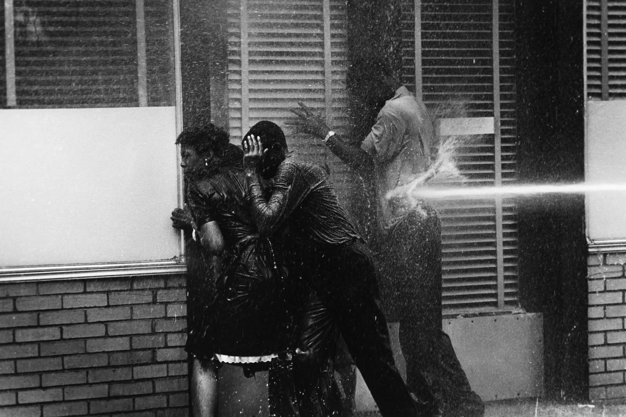 A black-and-white photograph of 3 people, leaning against the glass windows of a brick building, being sprayed with high-pressure fire hoses.