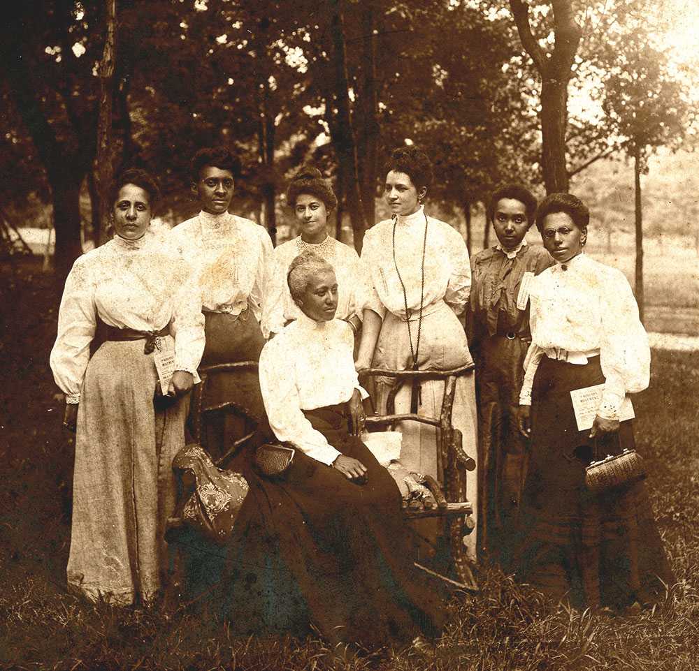 Black and white group portrait of seven women in an outdoor setting.  Six are standing and one is seated on a bench.