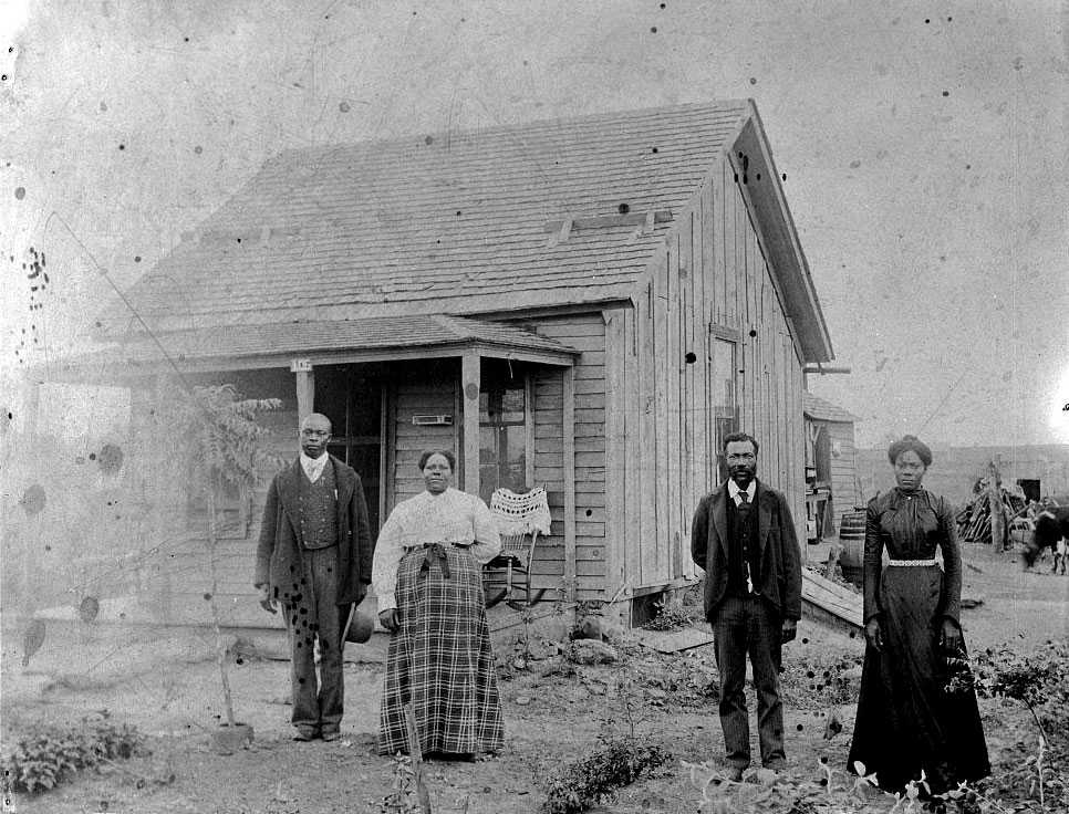 Two couples stand outside a small wooden housen. The land has various plants around the building.