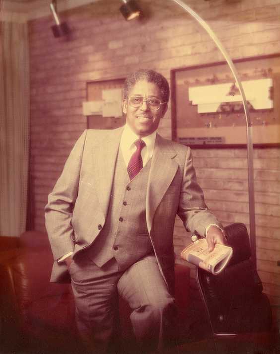 Architect John Chase wearing a three-piece suit and glasses, holding a rolled-up newspaper while leaning against a chair. The background includes brick walls with framed items and lighting fixtures, suggesting a formal or office setting.