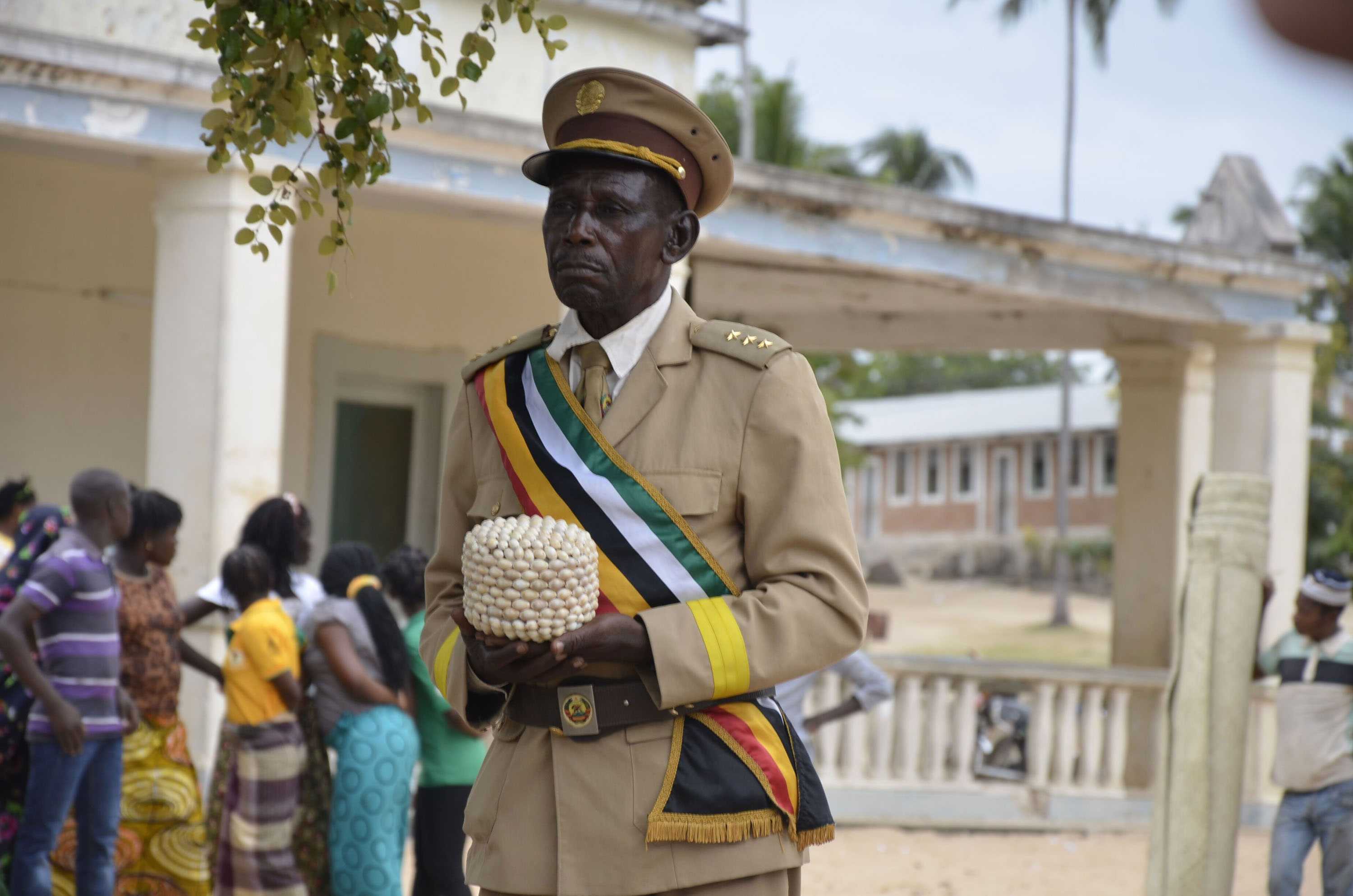 A stotic older man dressed in a traditional uniform holding a cowrie shell basket.