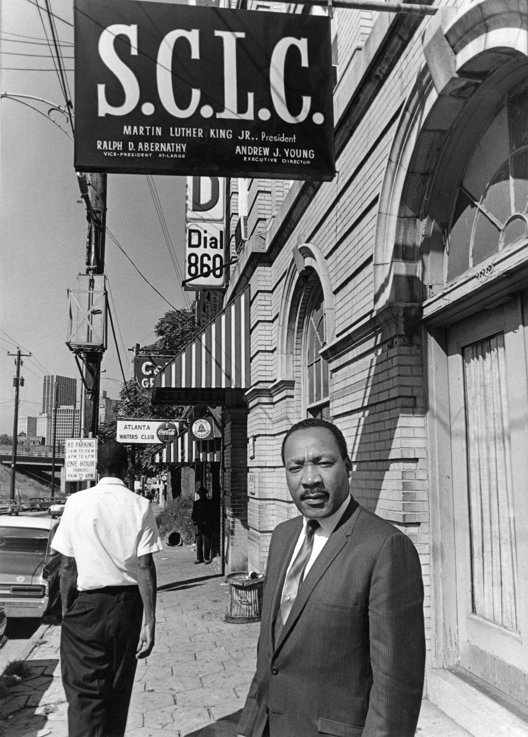 In a black suit, Martin Luther King Jr. stands outside on the sidewalk of the SCLC Headquarters in Atlanta.