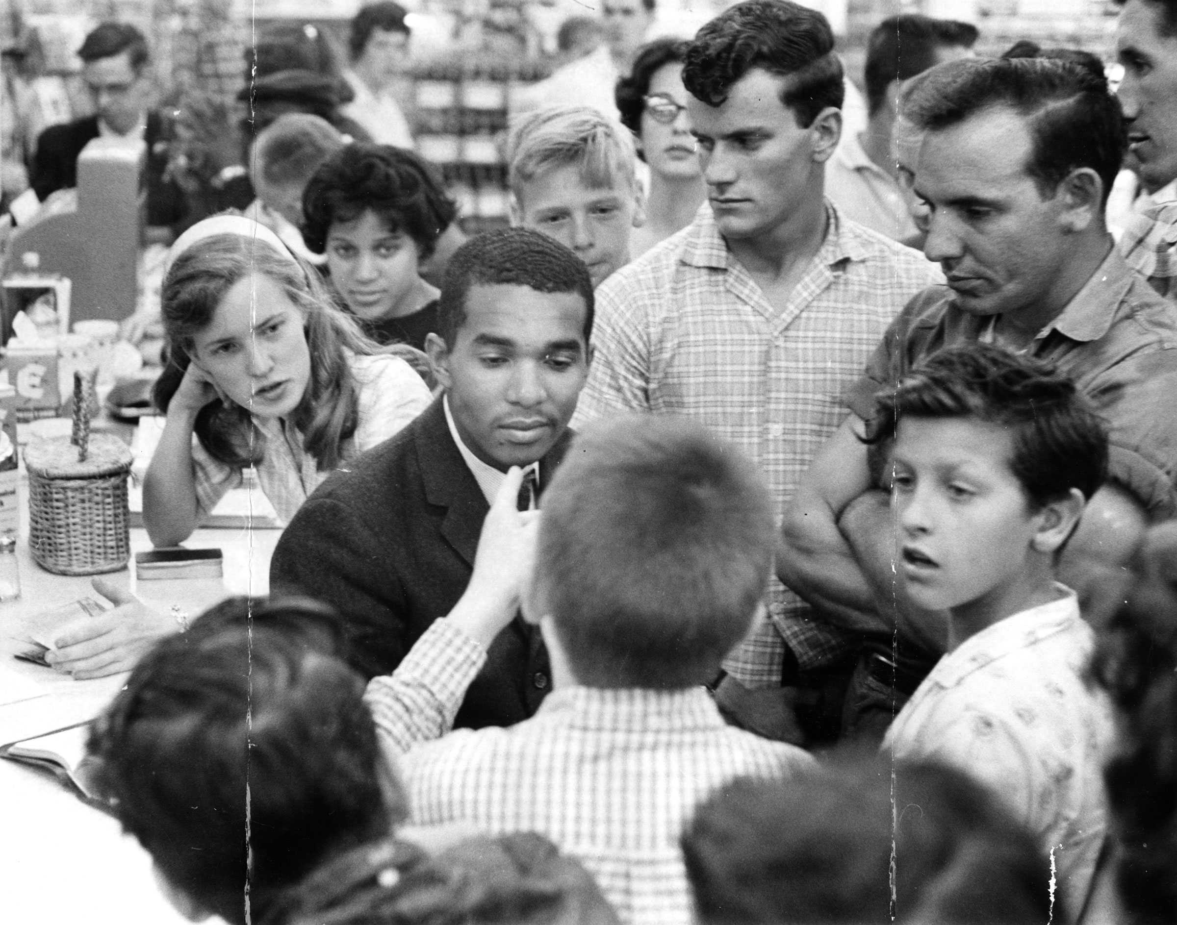 A Black college student sits at a counter with a crowd of white people surround him. A younger boy is angrily pointing at the student.