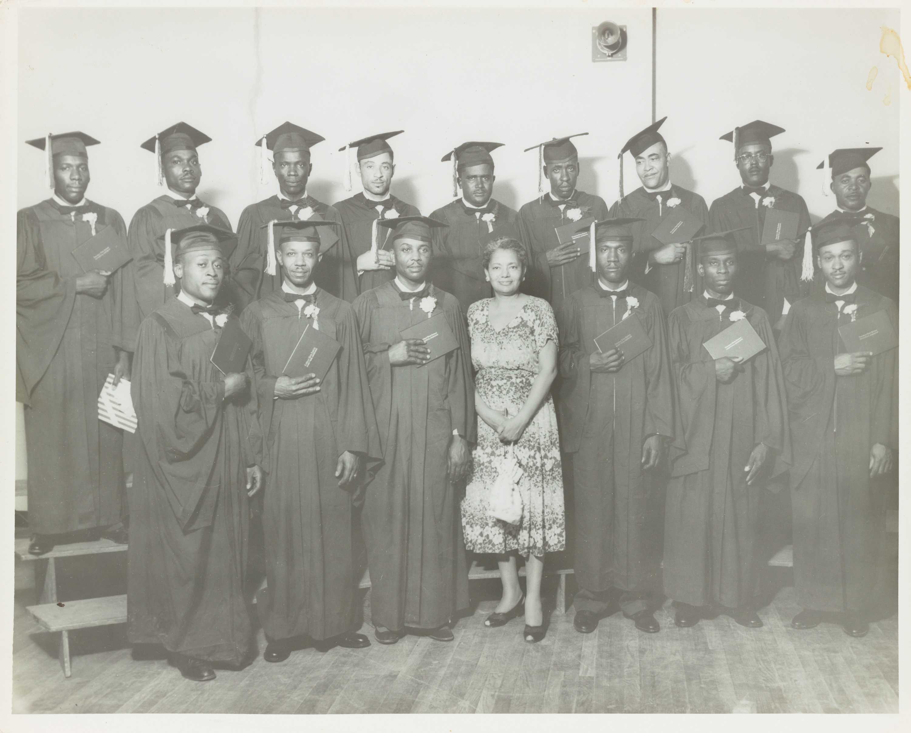 Group portrait of young men wearing graduation caps and gowns.  Each man holds his diploma to his chest with right hand.  In the middle of the first row an unidentified woman poses with them.