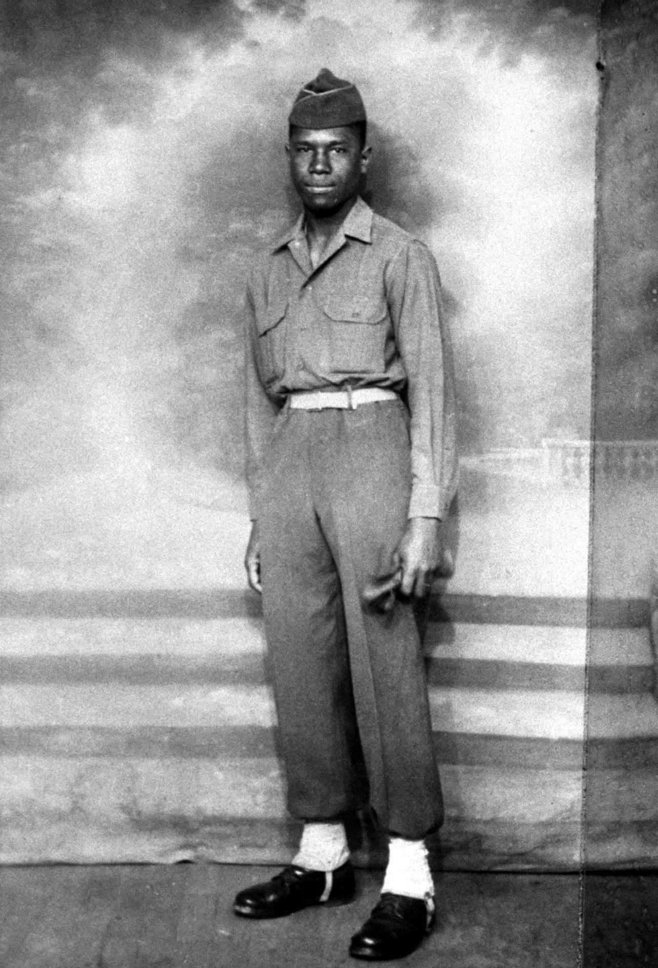 A young African American man in military uniform stands against a plain backdrop, wearing a buttoned shirt, high-waisted trousers, and a cap. He has a serious expression and is looking directly at the camera.