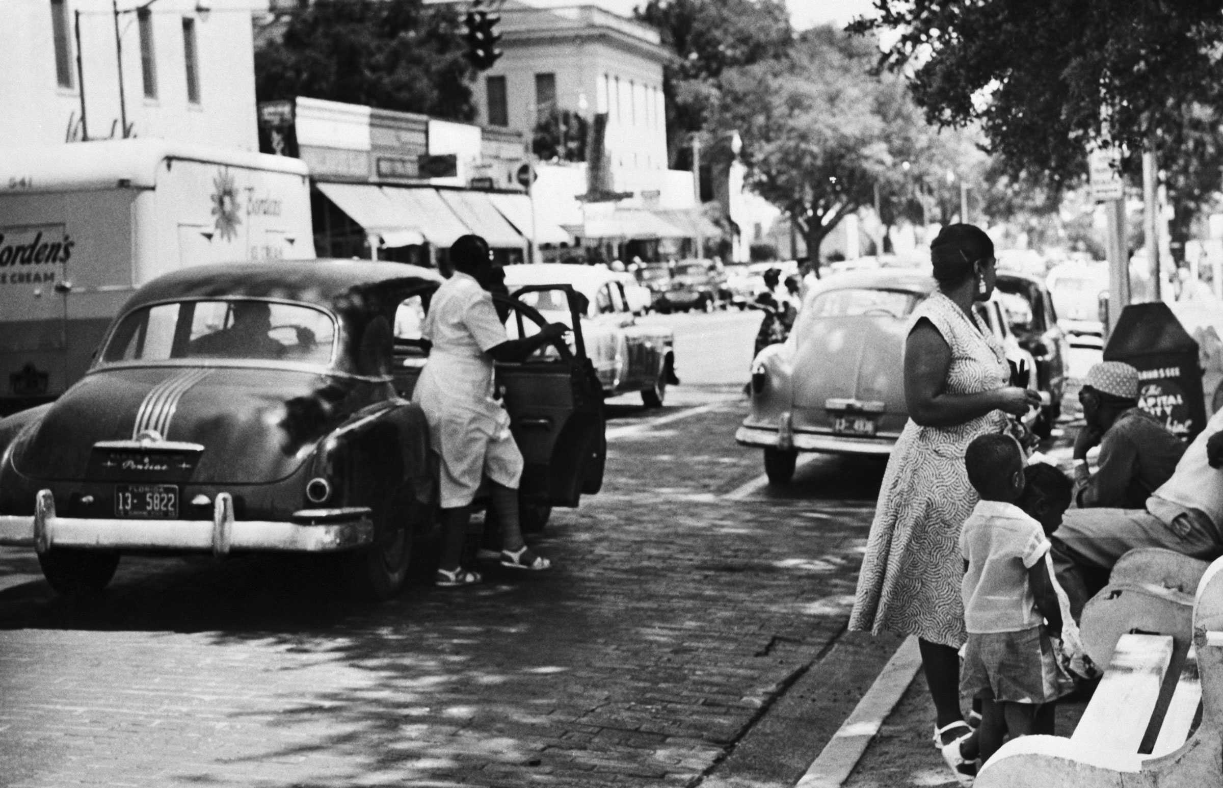 A black and white photograph of boycotters waiting and get out of carpools. A family waits by a bench on the sidewalk.