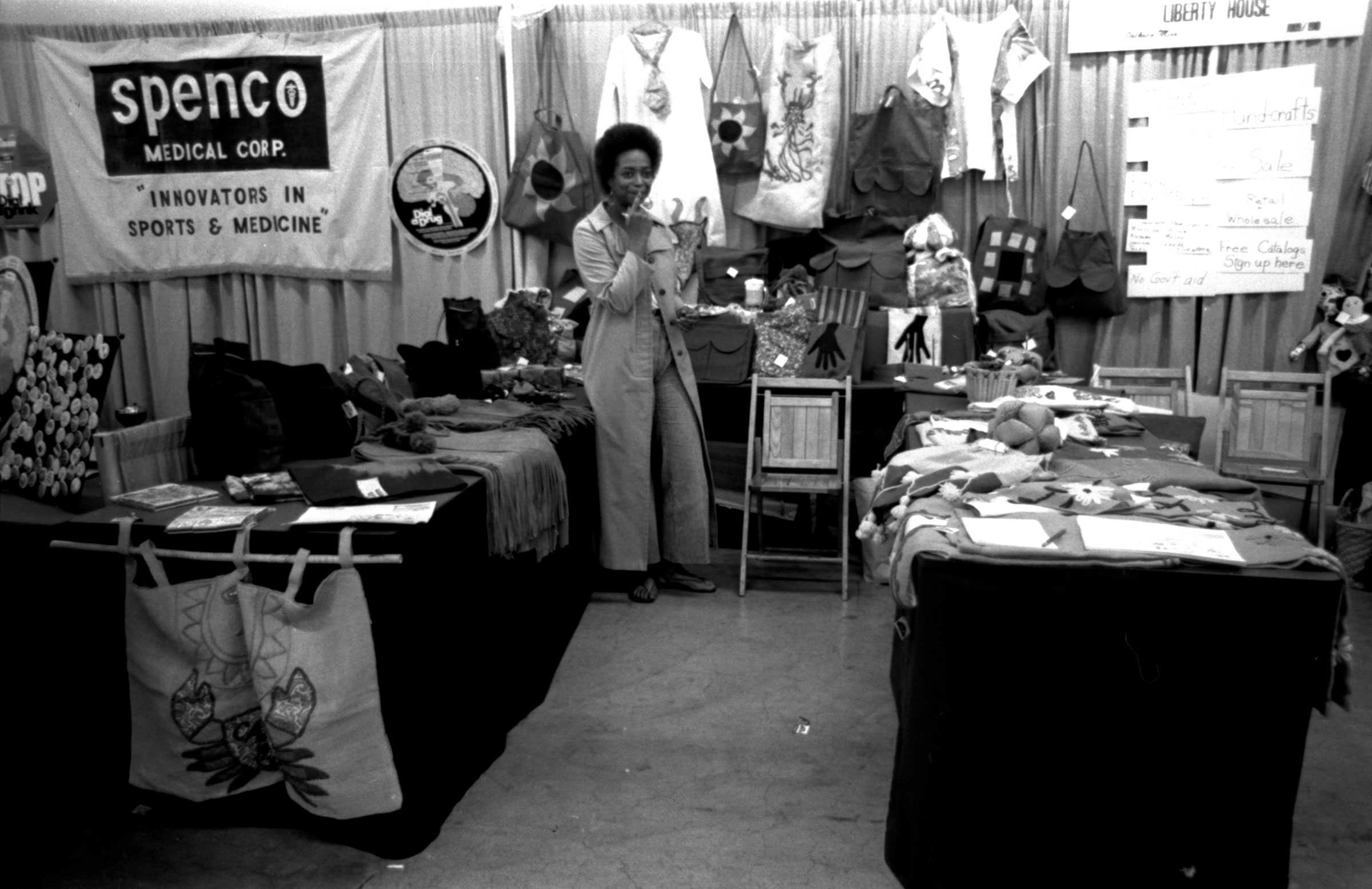 Black-and-white photo of Rep. Fay Bellamy Powell at a Los Angeles Trade Show, standing in a booth. A sign reads "Spenco Medical Corp.", with textiles and items displayed.